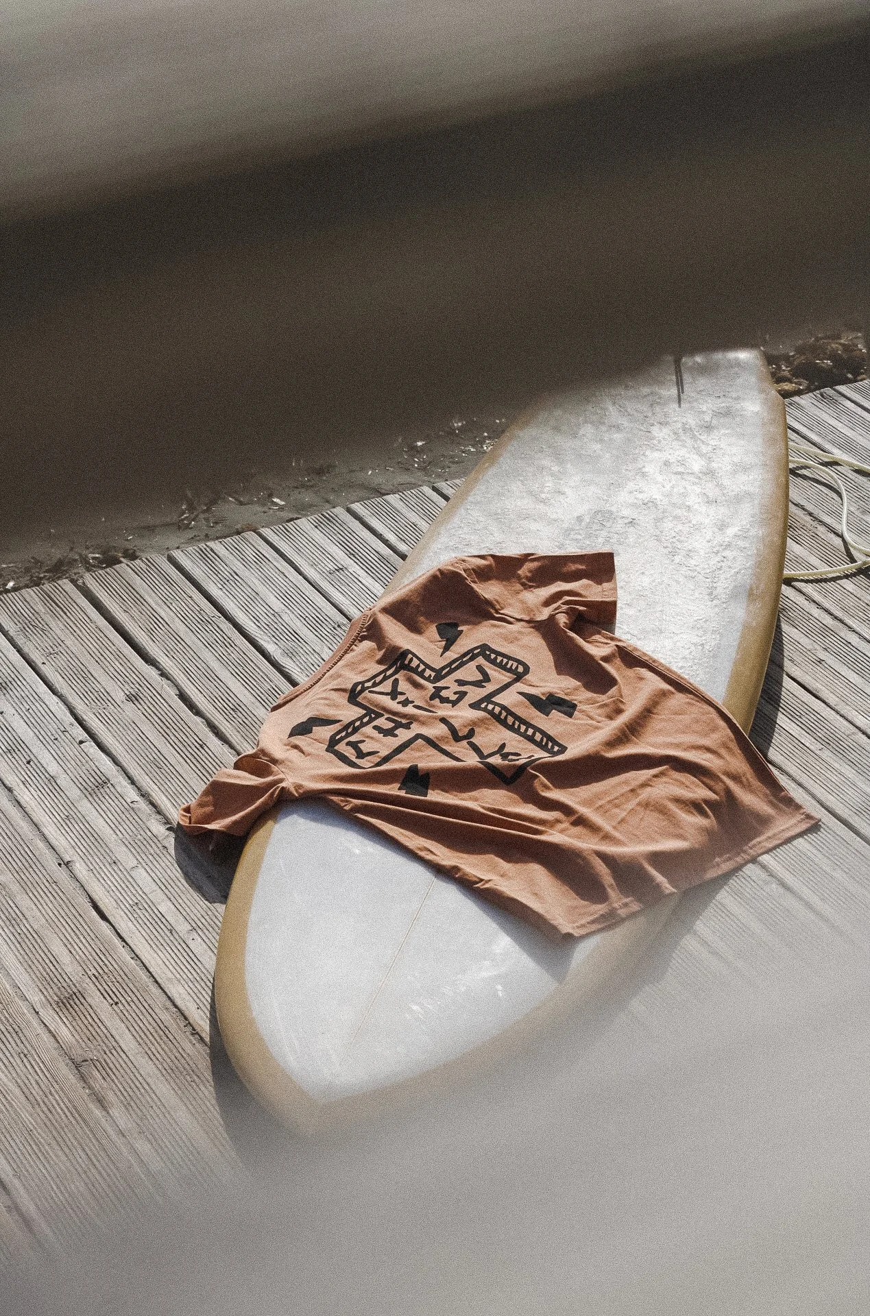Une planche de surf beige sur une plateforme en bois, avec un t-shirt marron aux motifs noirs posé dessus.