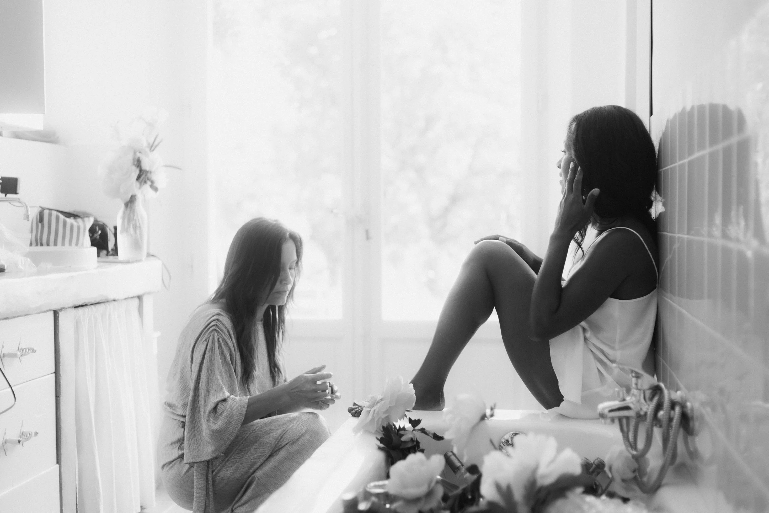 Deux femmes dans une salle de bain, l'une assise dans une baignoire, l'autre assise par terre, discutant ou partageant un moment intime.