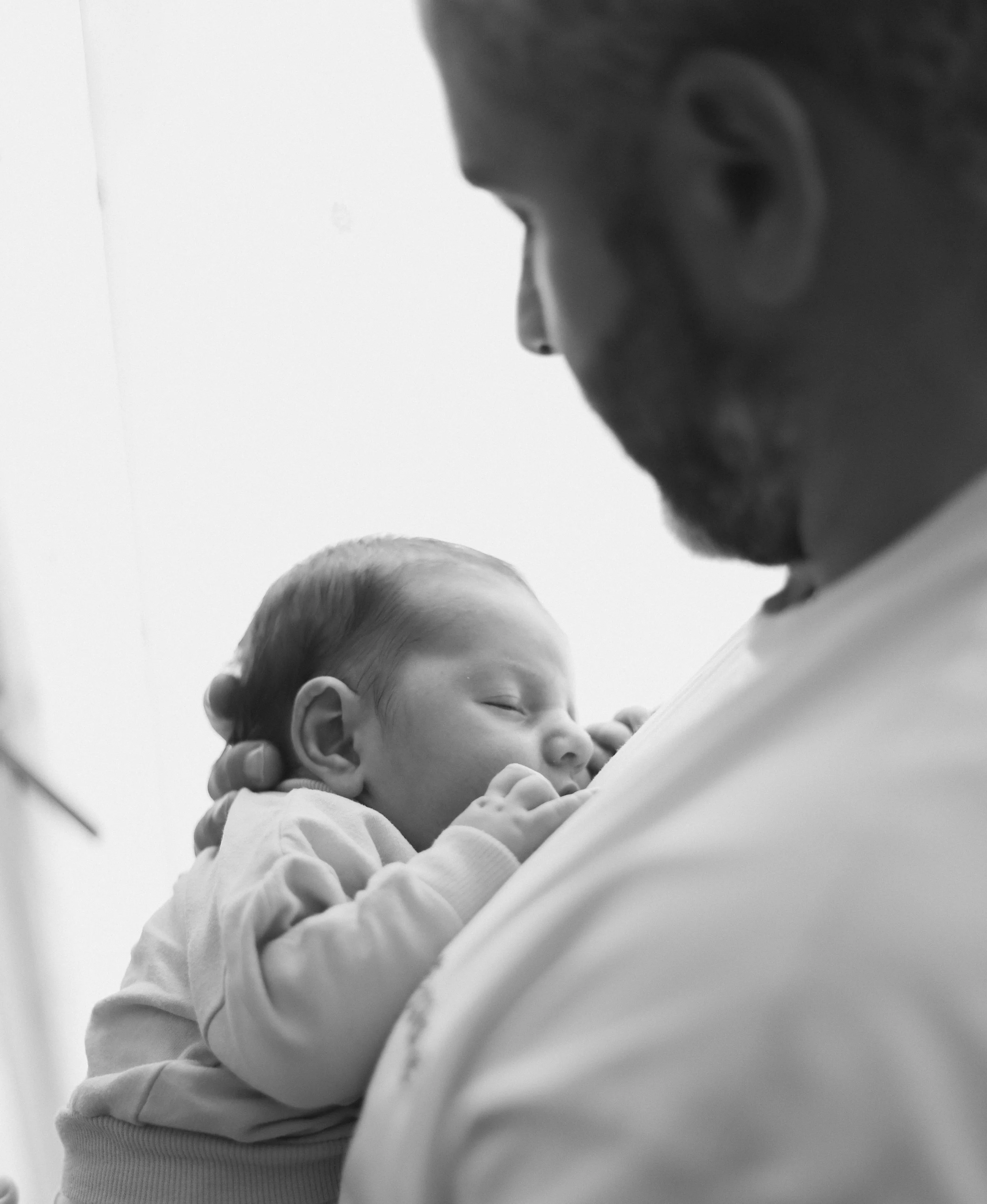 Un adulte tient un bébé endormi contre lui, dans un environnement intérieur, en noir et blanc.