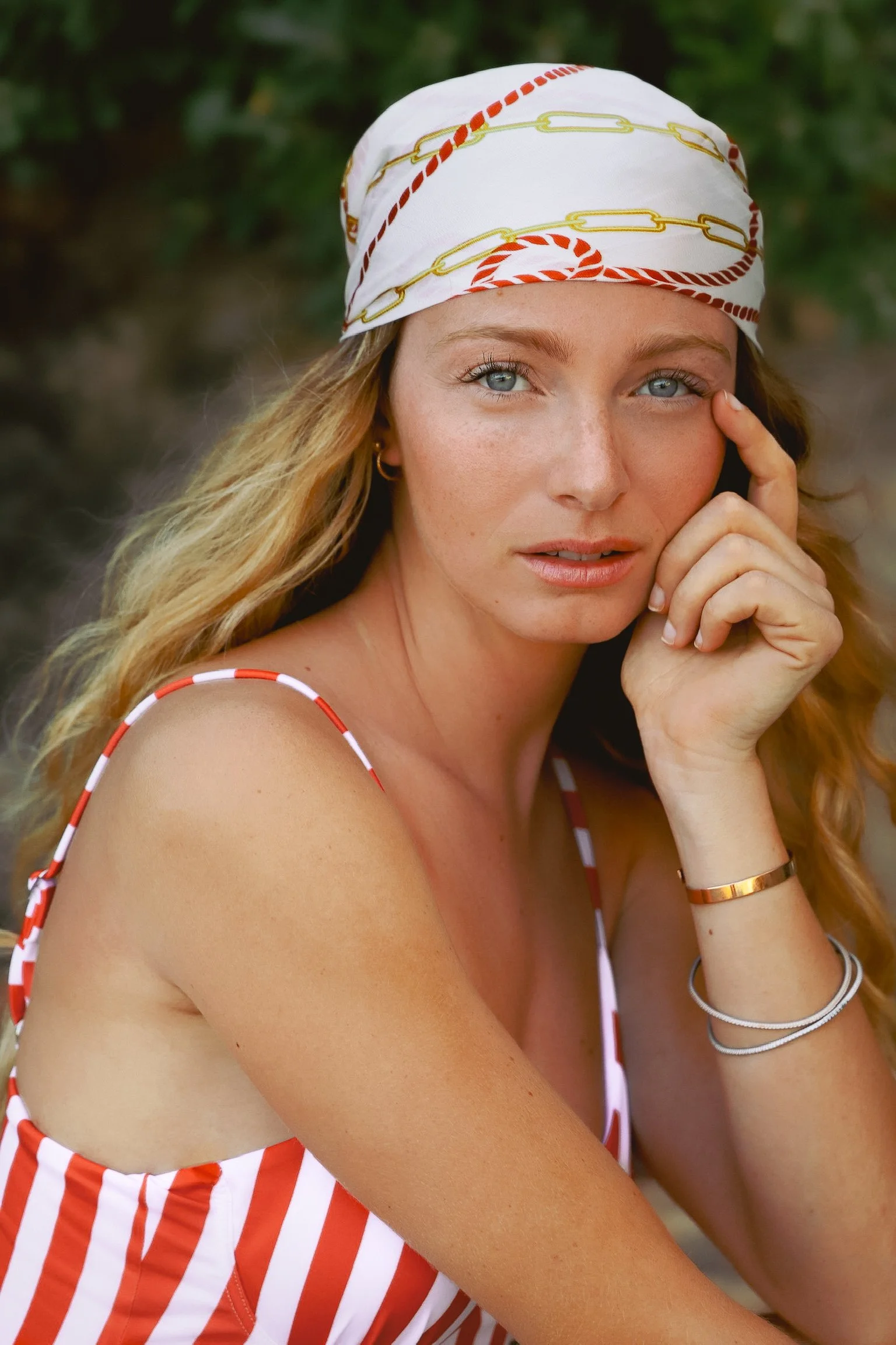 Une femme portant un turban blanc avec des motifs rouge et jaune, un maillot à rayures rouges et blanches, et plusieurs bracelets sur le poignet gauche, posant devant un fond nature.