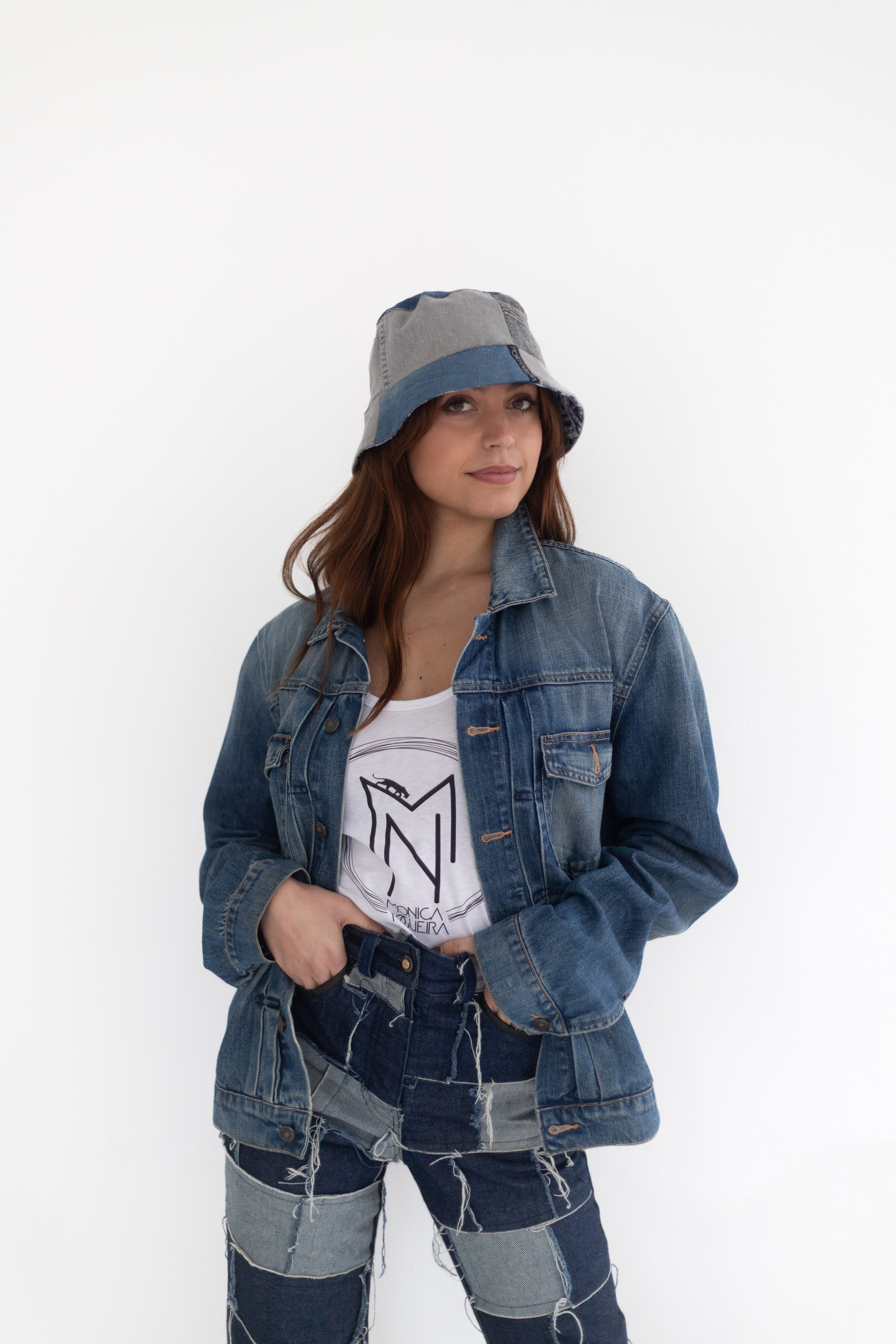 Une jeune femme avec des cheveux bruns portant un chapeau en denim, une veste en jean, et un t-shirt blanc avec un logo, posant contre un fond blanc.