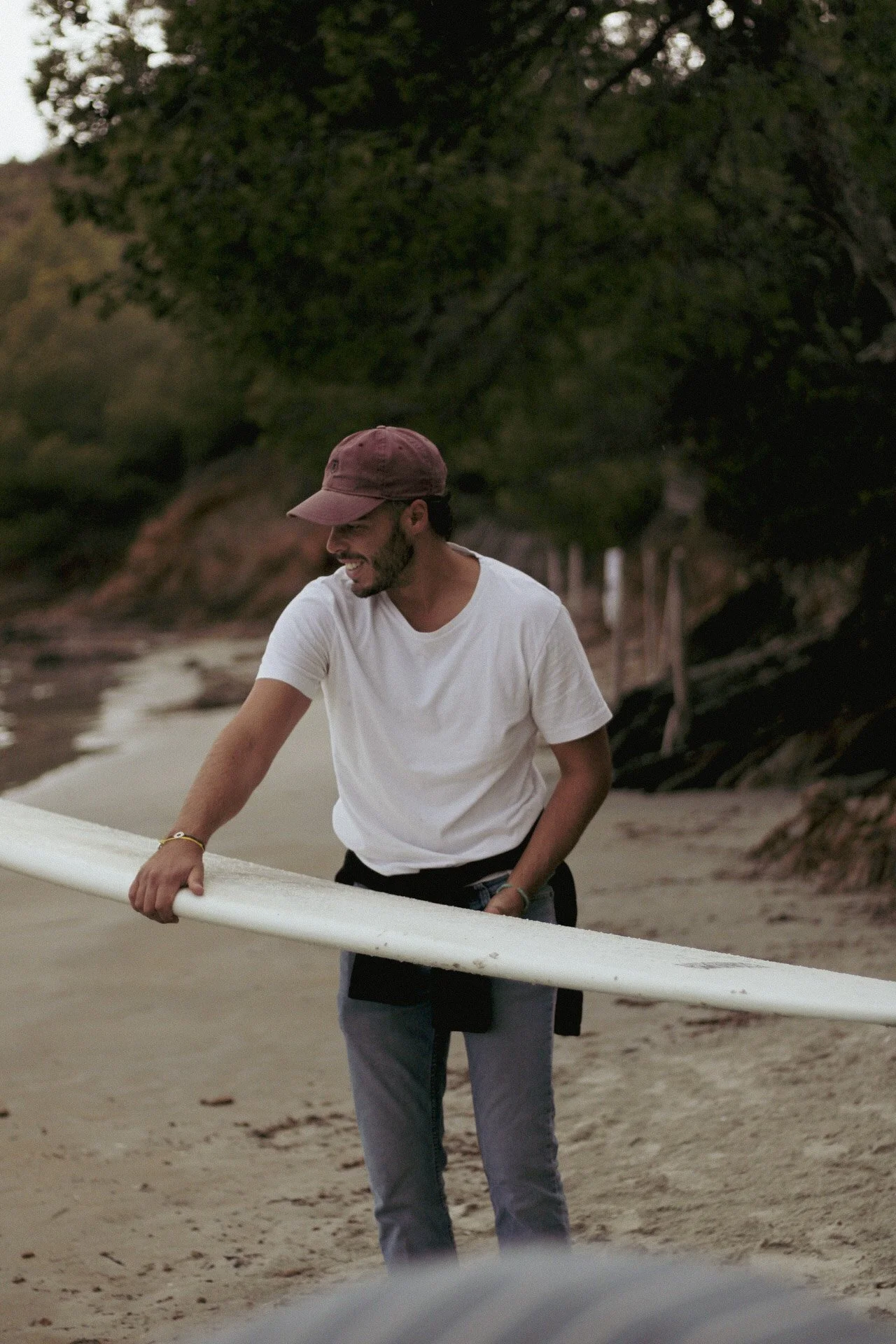 Jeune homme souriant, portant un chapeau marron et une chemise blanche, tenant une planche de surf sur la plage