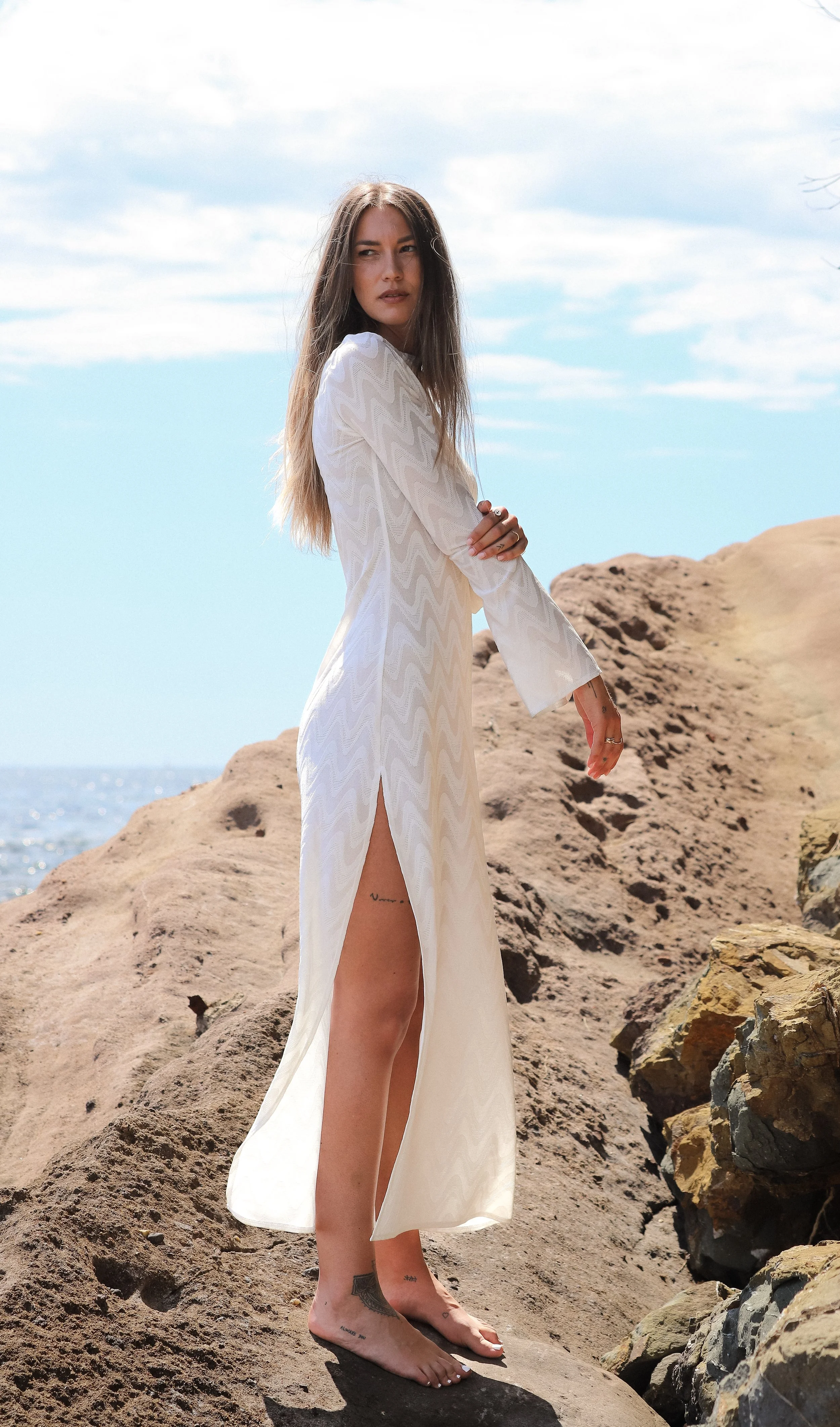 Femme debout sur des rochers au bord de la mer, portant une robe longue blanche avec une fente, sous un ciel partiellement nuageux.