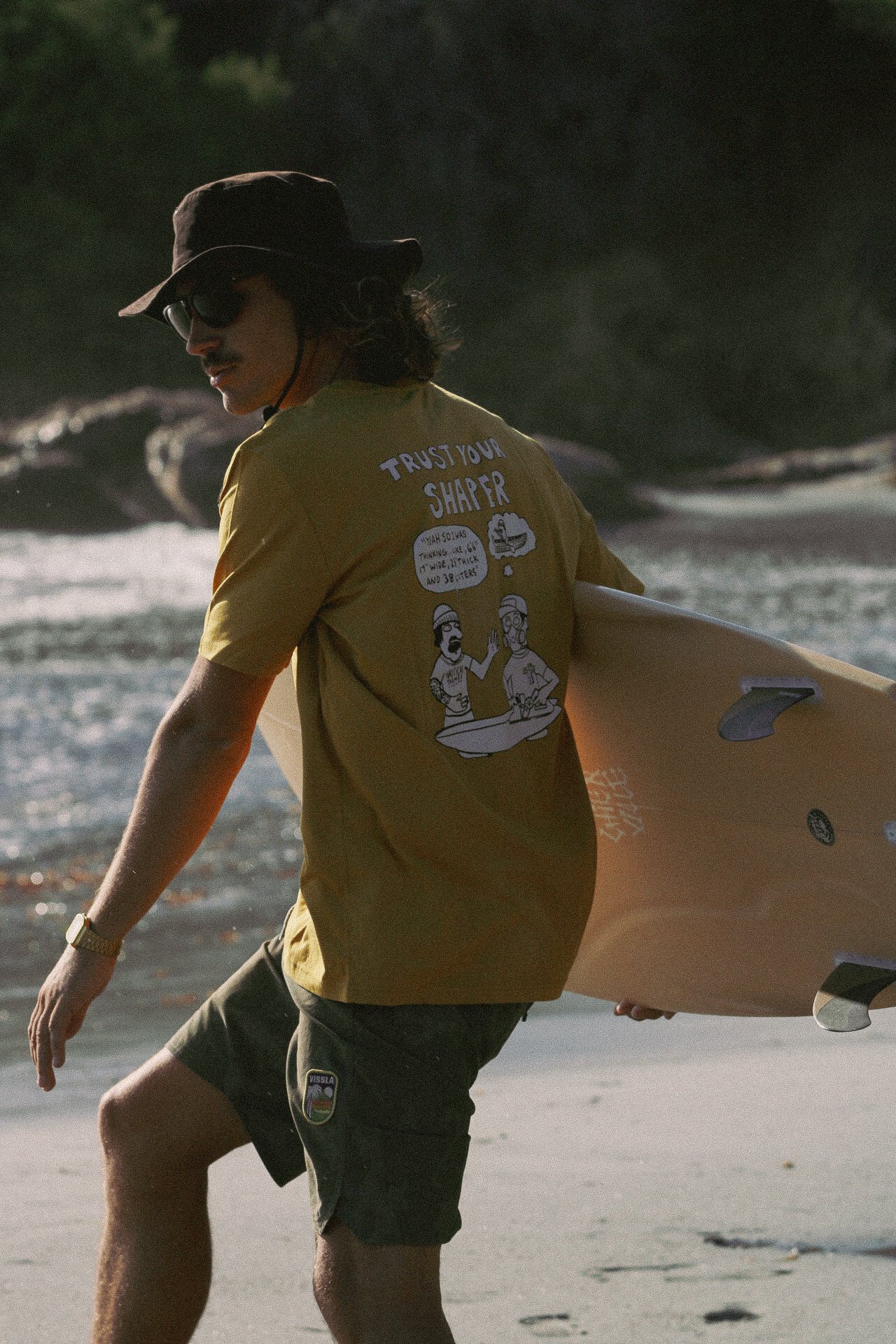 Un homme portant un chapeau, des lunettes de soleil, un t-shirt jaune avec un dessin humoristique et un texte, tient une planche de surf sur une plage au coucher du soleil.
