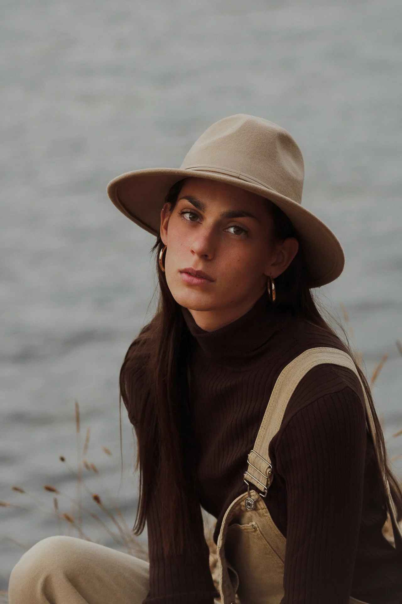 Jeune femme portant un chapeau beige, une écharpe beige, un pull en maille marron et un pantalon beige, assise près d'un lac lors d'une journée grise.