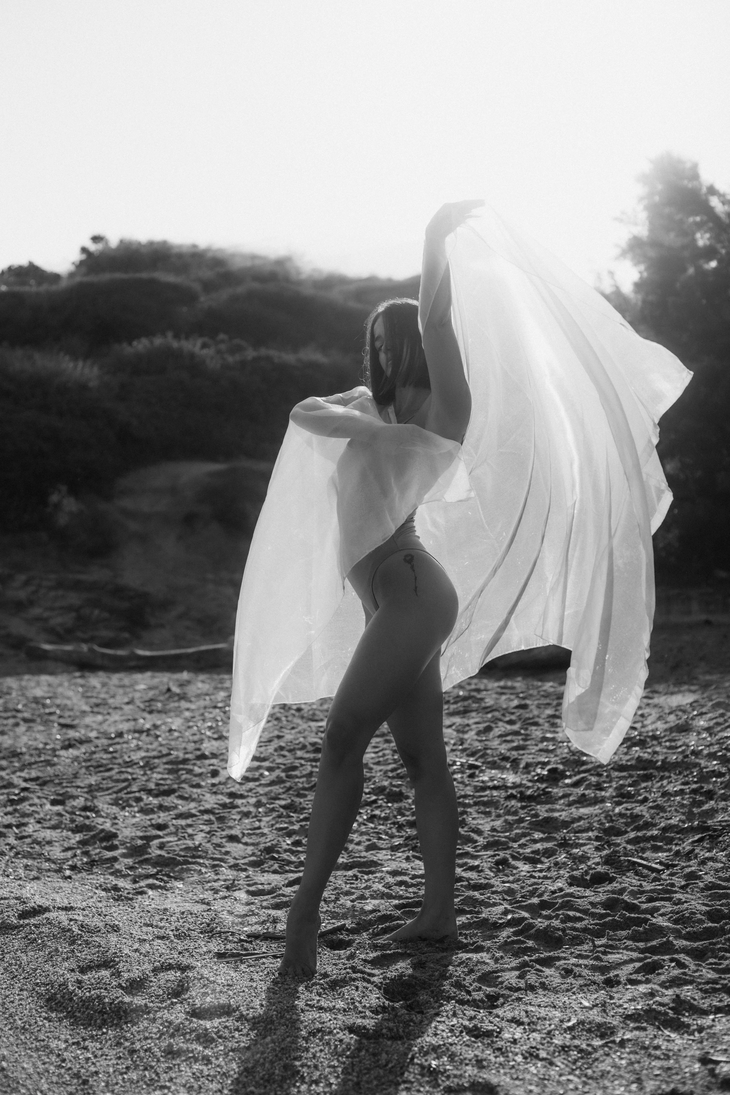 Une femme nue tenant un tissu léger sur une plage au coucher du soleil, en noir et blanc.