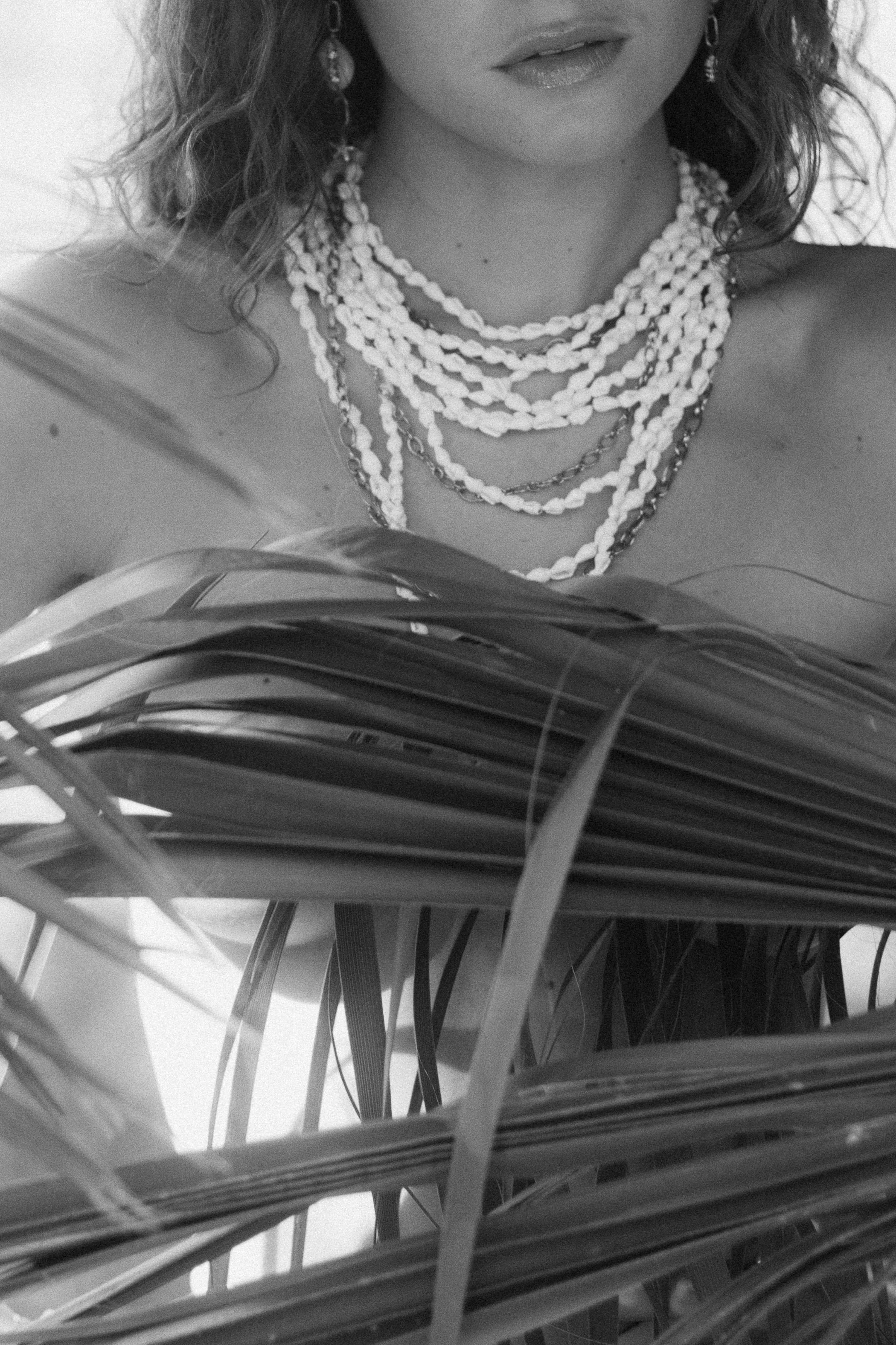 Une femme porte plusieurs colliers de perles et des boucles d'oreilles avec des cheveux bouclés, la moitié inférieure de son visage est visible, et elle est partiellement cachée derrière de grandes feuilles.