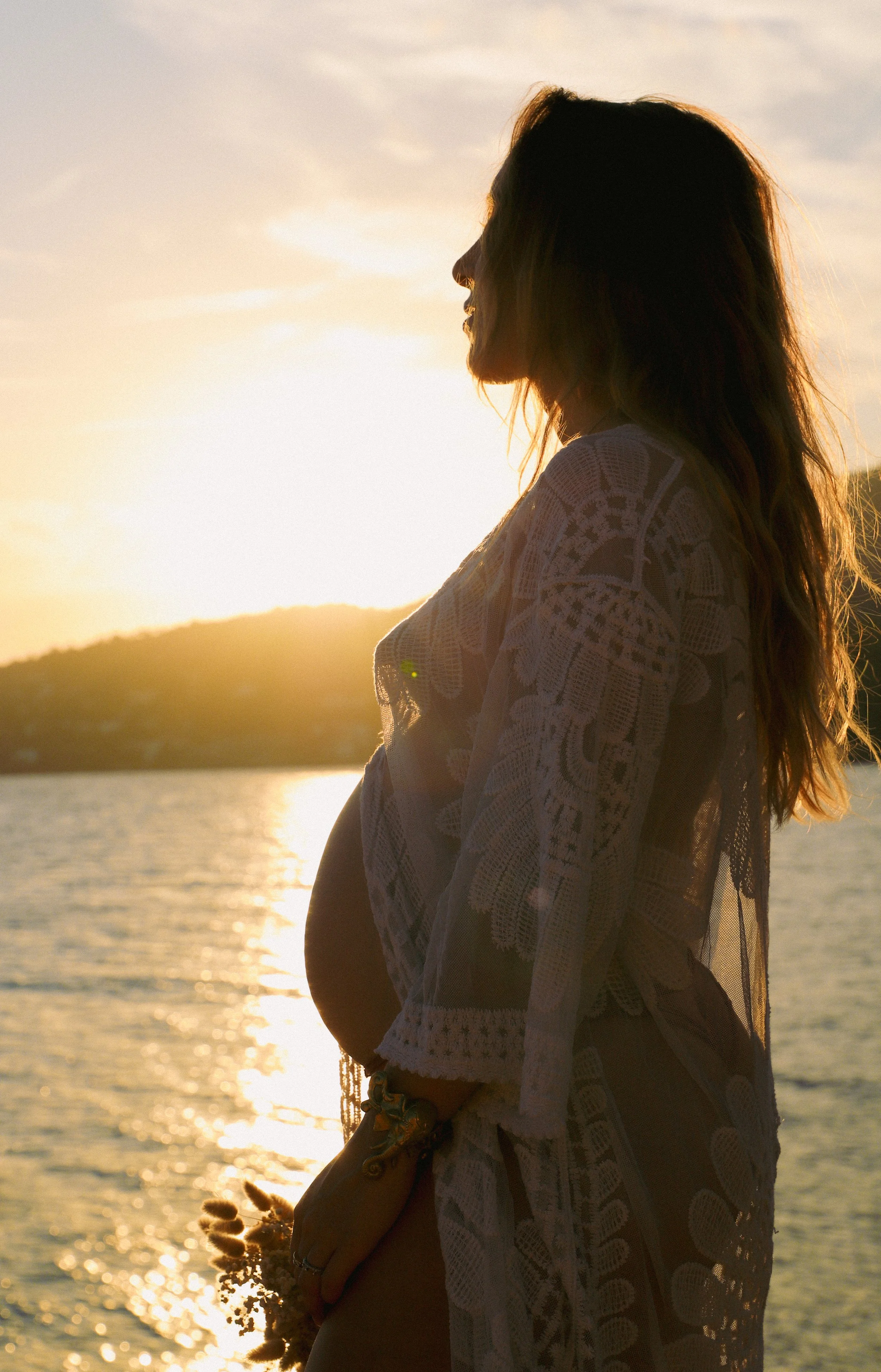 Femme enceinte en silhouette portant une robe en dentelle, se tenant le ventre au bord d'une rivière au coucher du soleil.