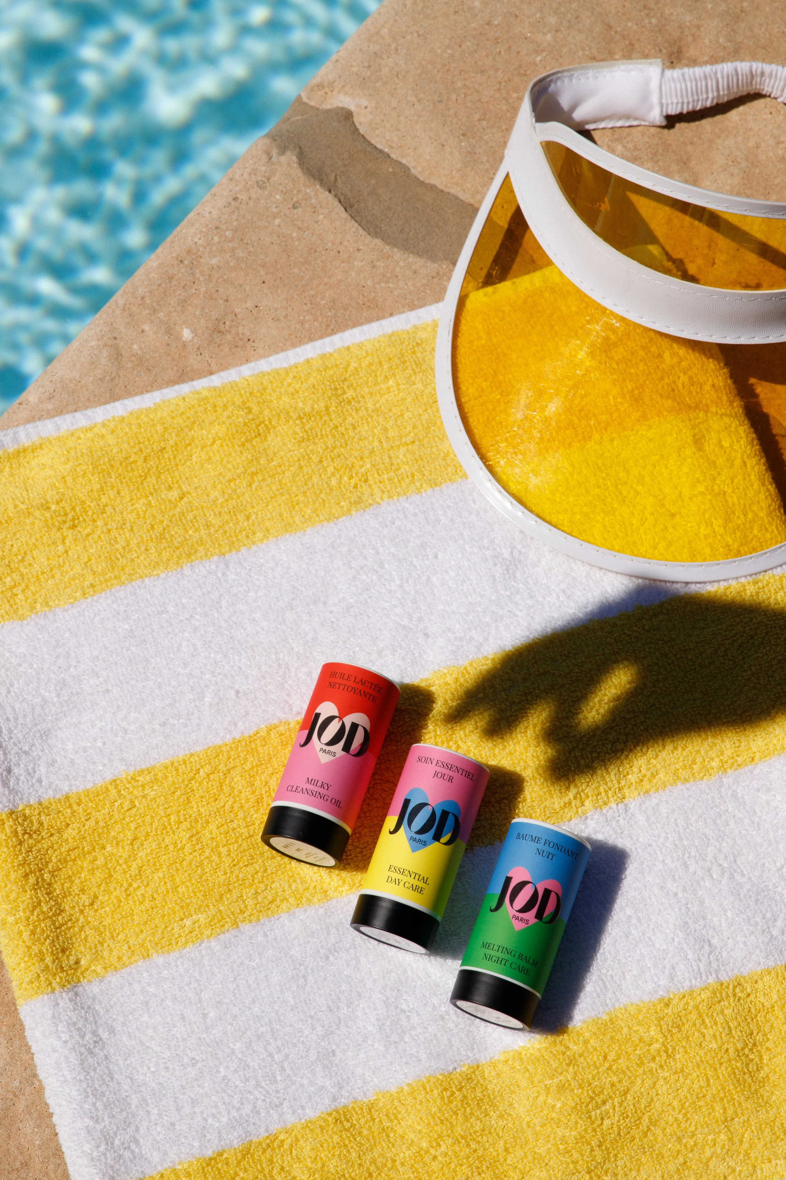 Crème solaire de différentes couleurs sur une serviette de plage, avec une piscine en arrière-plan et un chapeau de soleil.