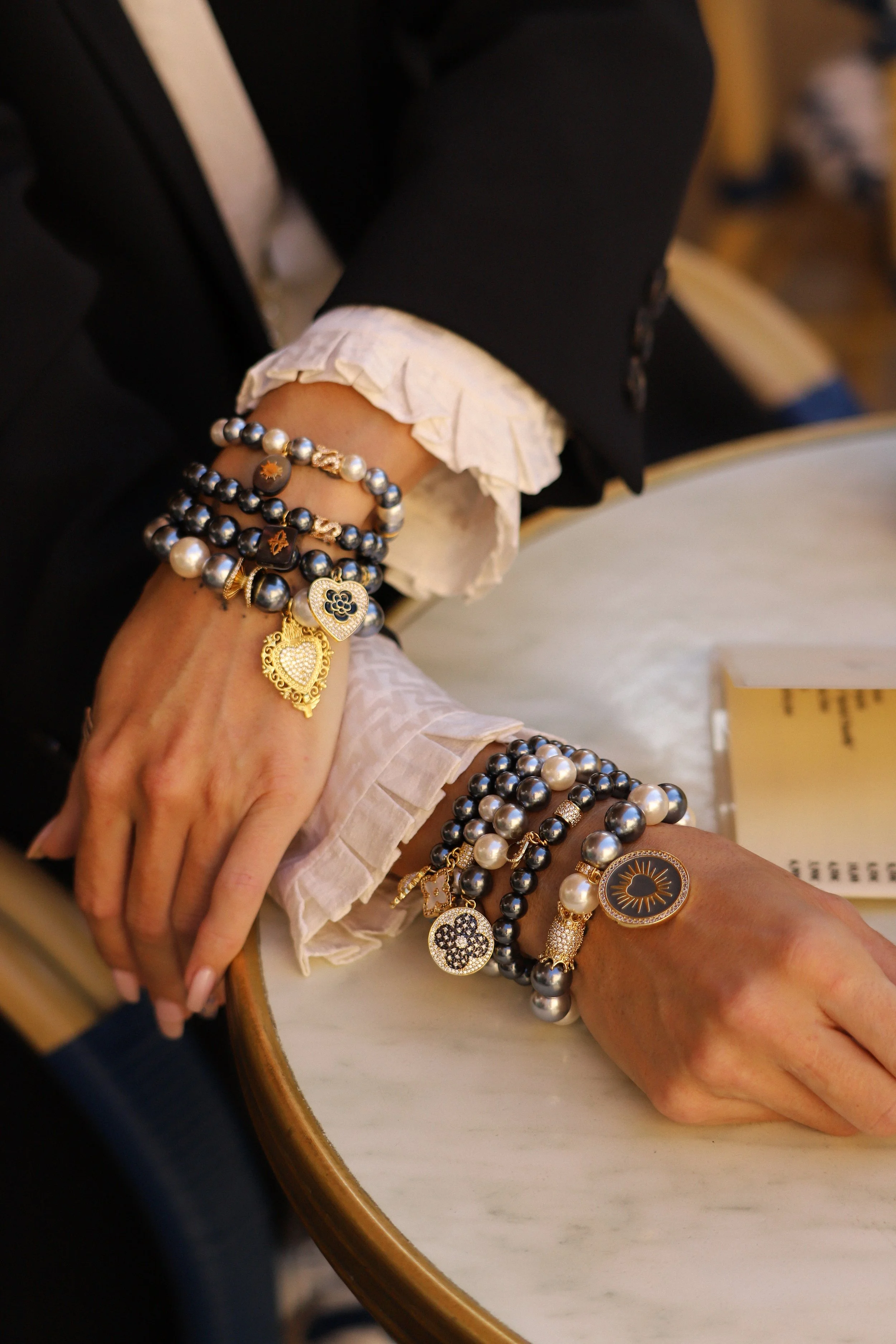 Poignet d'une femme avec plusieurs bracelets en perles noires, blanches et ornées de charms, portant une manche blanche froissée et une veste noire, posé sur une table blanche.