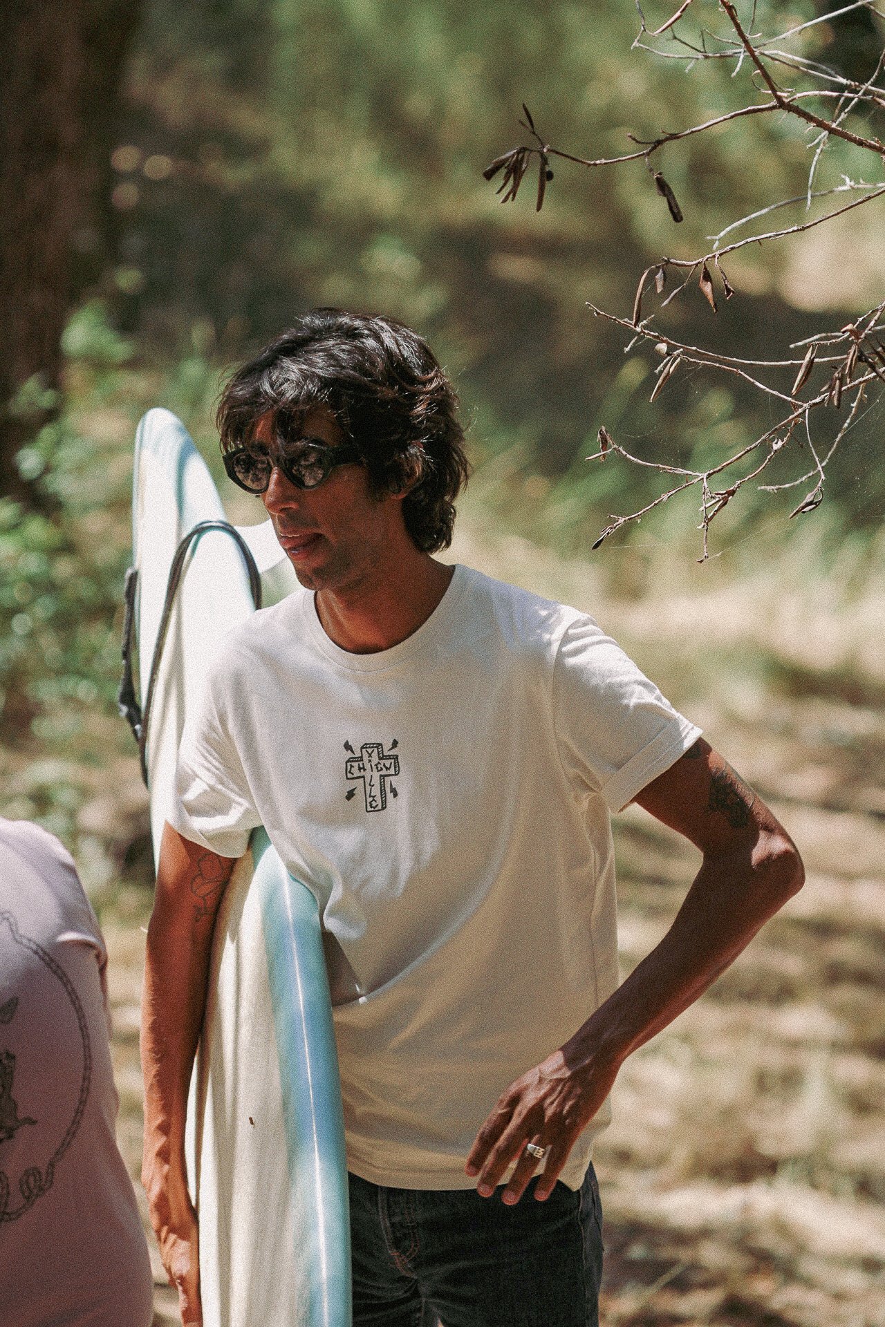 Un homme avec des cheveux longs, portant des lunettes de soleil, un t-shirt blanc avec un dessin de croix, tient une planche de surf dans un environnement naturel avec des arbres et des branches.