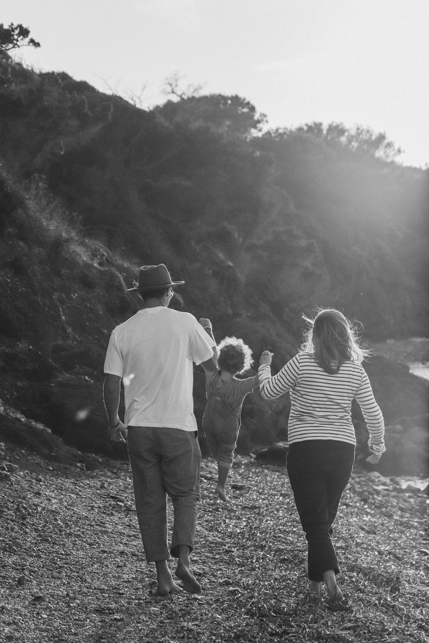 Une famille marche pieds nus sur la plage au coucher du soleil, vue de dos, en noir et blanc.