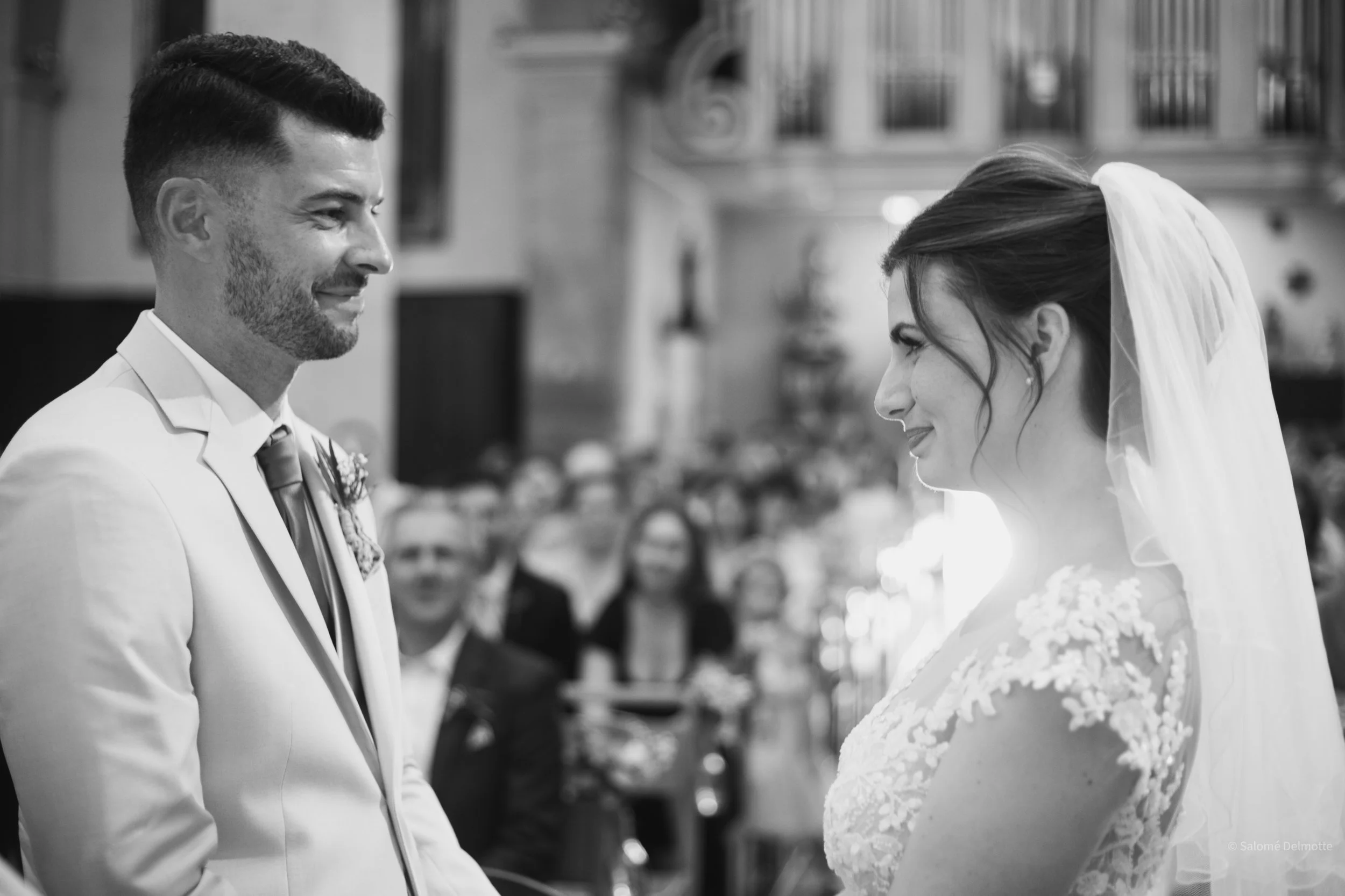 Un couple de mariés échangeant leur vœux lors de leur mariage dans une église, avec des invités en arrière-plan.