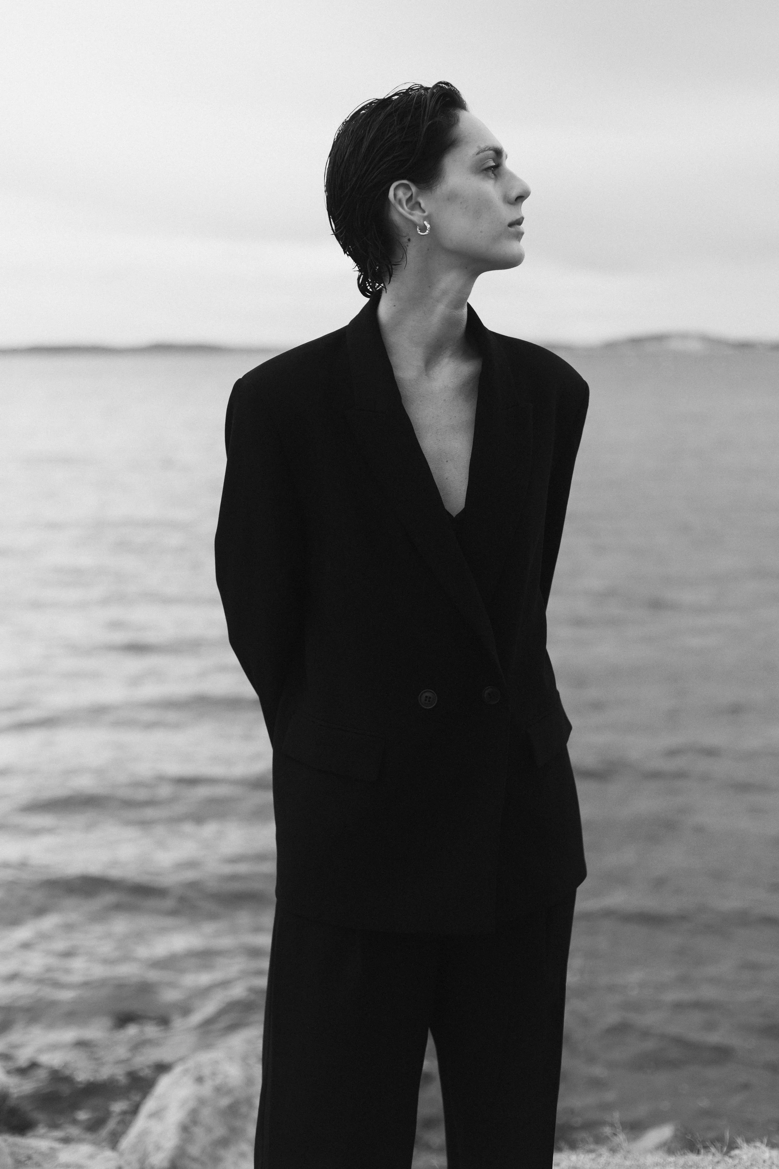 Une femme en costume noir regarde vers la droite, debout près d'un corps d'eau, en noir et blanc.