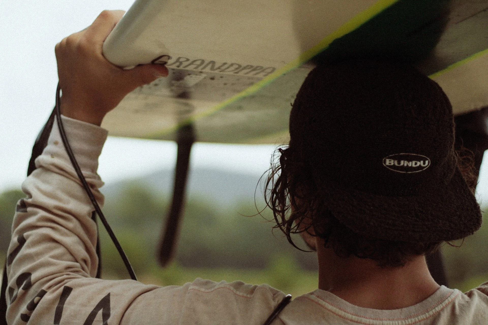 Une personne porte un bonnet noir avec le logo 'BUNDO' et tient une planche de surf au-dessus de la tête dans un environnement naturel.