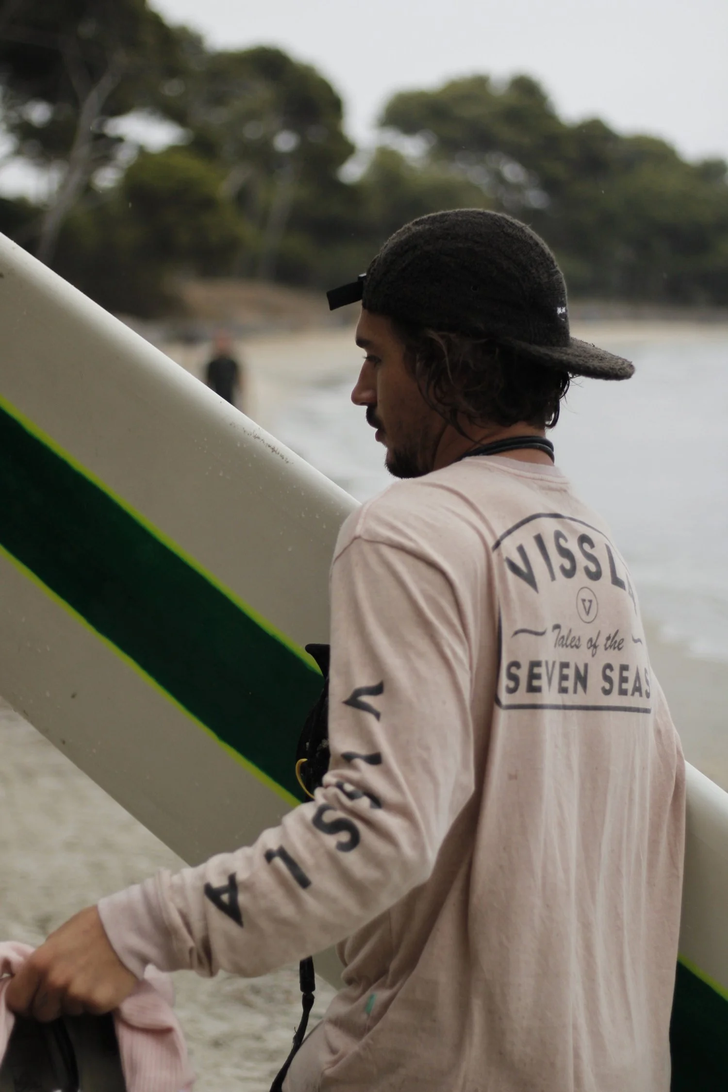 Jeune homme tenant une planche de surf sur une plage, portant un sweat-shirt beige avec écrit en anglais "VISSLA Tales of the SEVEN SEAS" et un chapeau noir, arrière-plan de végétation et de la mer.