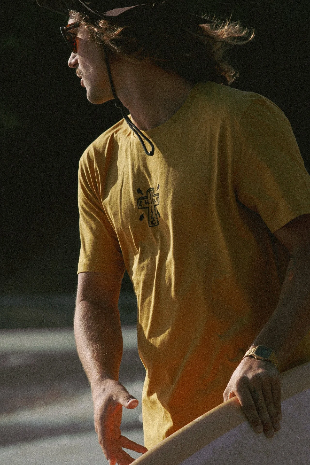 Un homme avec des cheveux longs portant un casque de vélo, une chemise jaune et un bracelet doré, tient une planche de surf sur la plage.