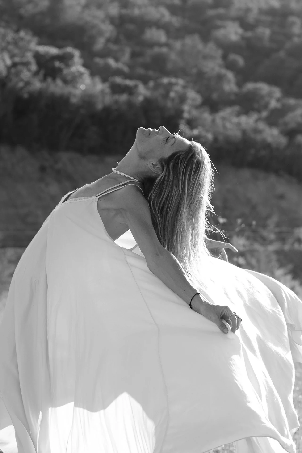 Une femme en robe blanche, assise sur une surface, en position de relaxation ou de méditation, avec une vue de paysage flou en arrière-plan, en noir et blanc.