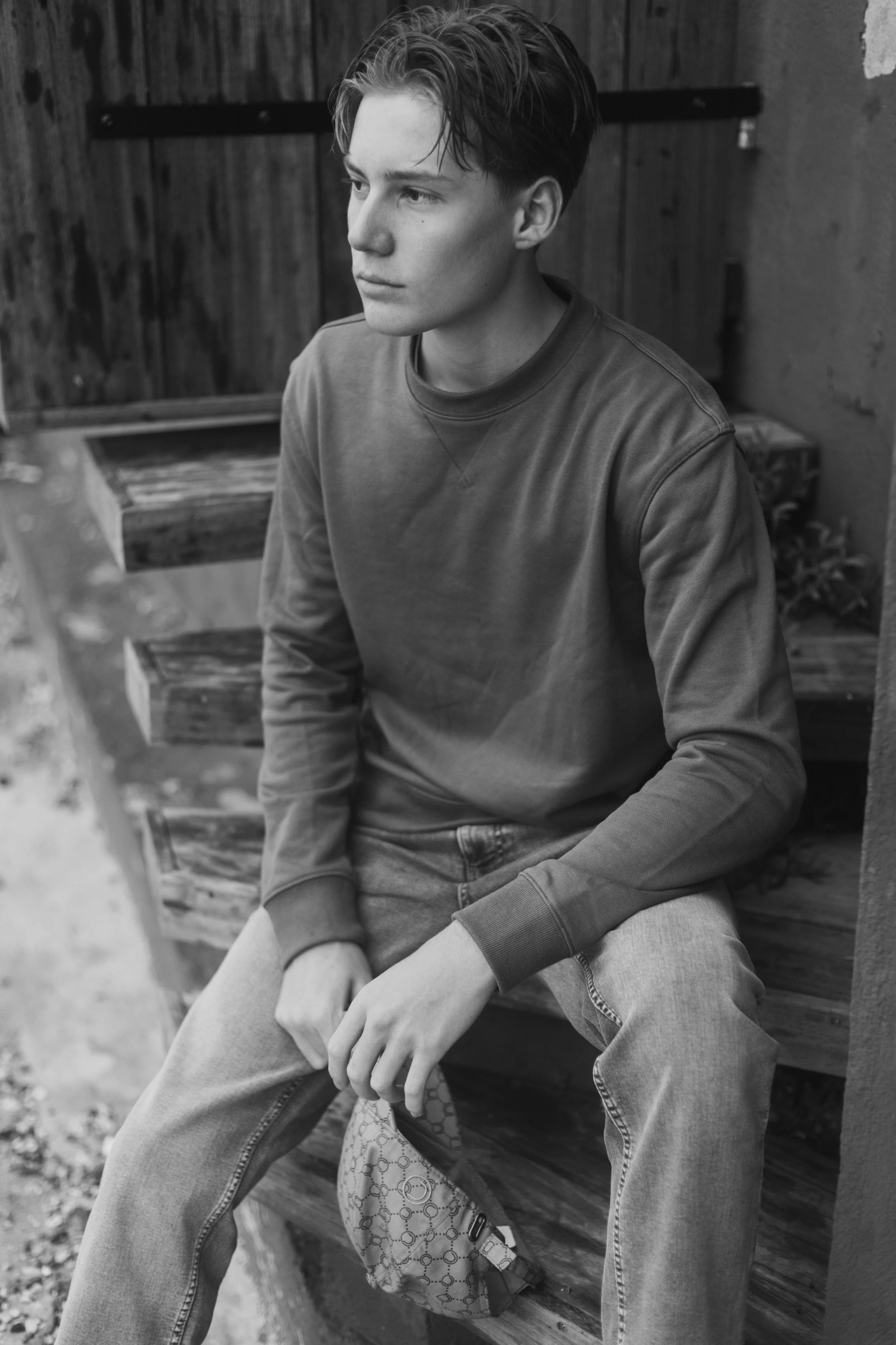 Jeune homme assis sur un banc en bois, regardant vers la gauche, portant un sweat à capuche et un jean, tenant un sac à, mesurant environ la hauteur du genou, dans un environnement extérieur.
