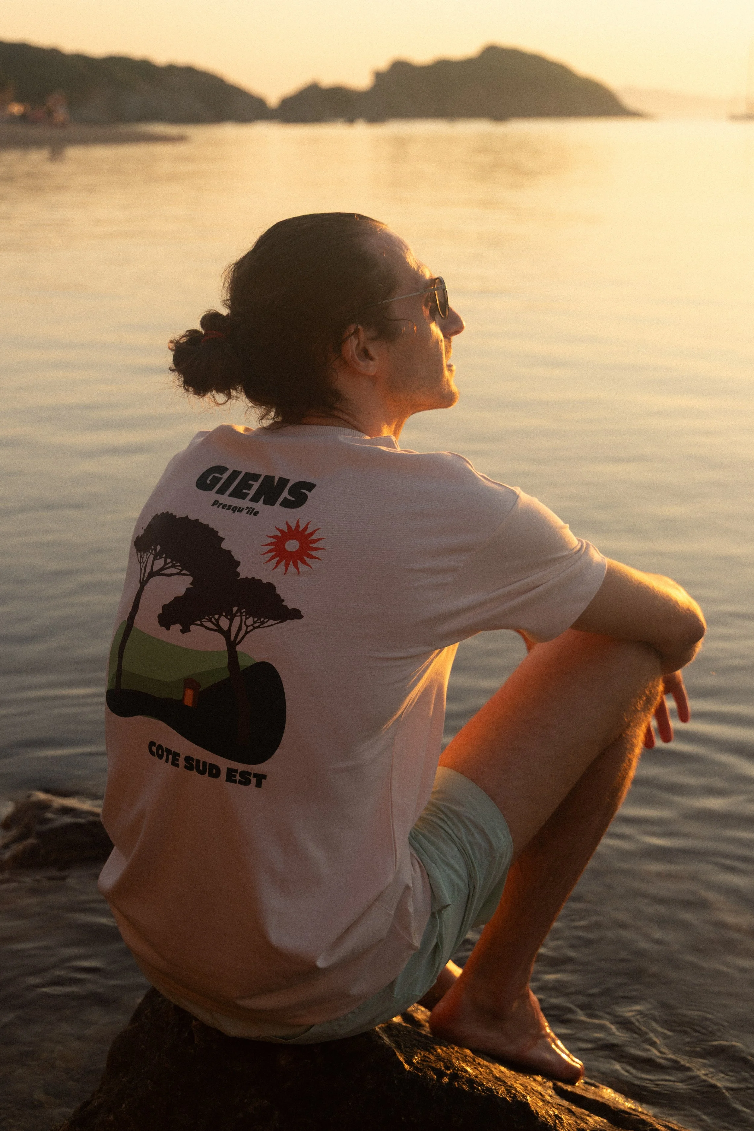 Un homme assis sur un rocher au bord de l'eau, regardant le coucher de soleil, portant un t-shirt blanc avec un dessin de deux arbres, un soleil rouge et le texte 'GIENS Presqu'île, Côté Sud Est'.