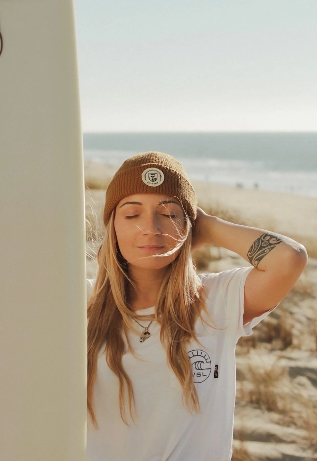 Jeune femme aux cheveux longs, portant un bonnet marron et une chemise blanche, se tenant à côté d'une planche de surf sur une plage ensoleillée, avec l'océan en arrière-plan.