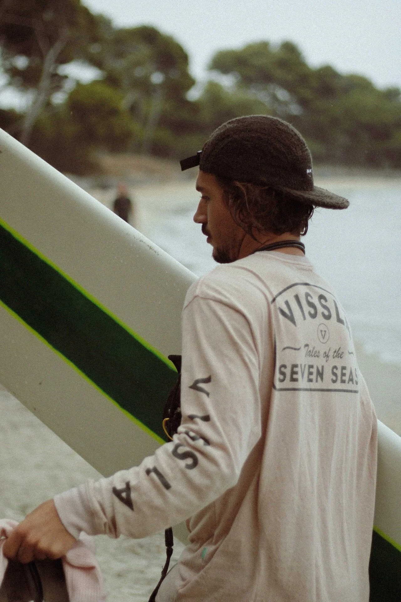 Un homme portant un sweat-shirt avec l'inscription 'VISSLA Tales of the SEVEN SEAS' et un chapeau, tenant une planche de surf sur une plage.