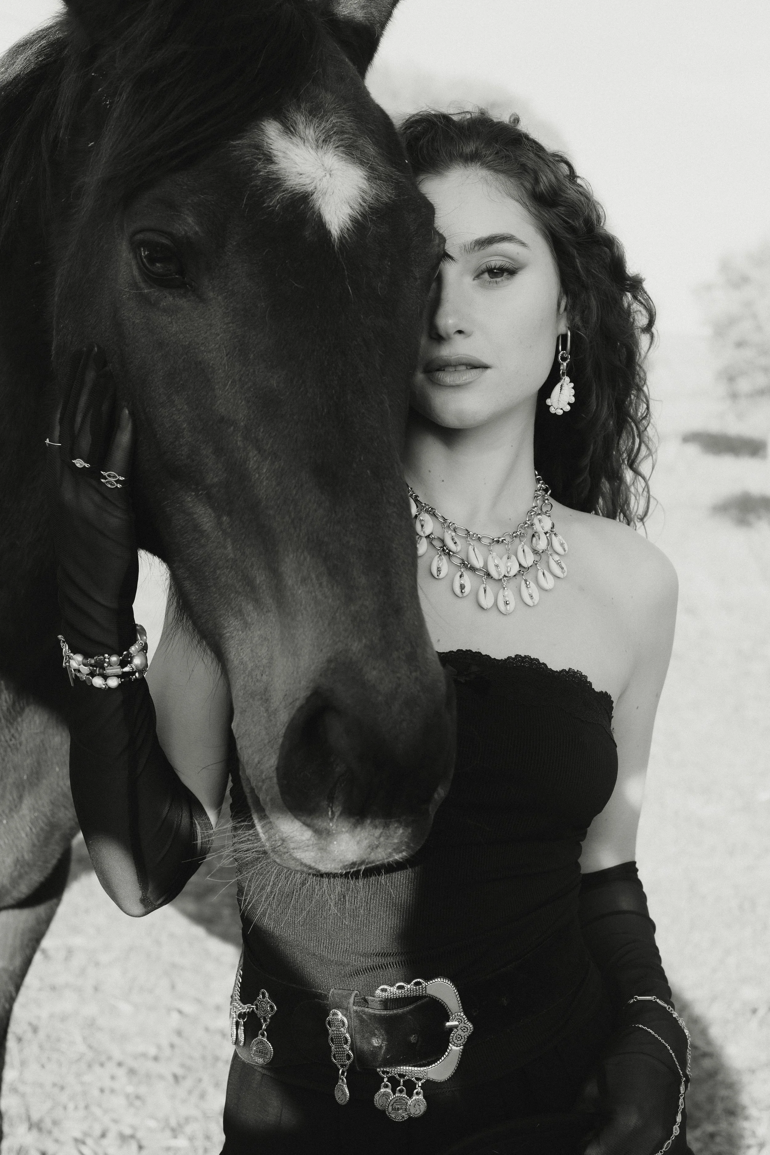 Une femme avec des bijoux élégants pose avec un cheval noir dans un paysage extérieur, en noir et blanc.