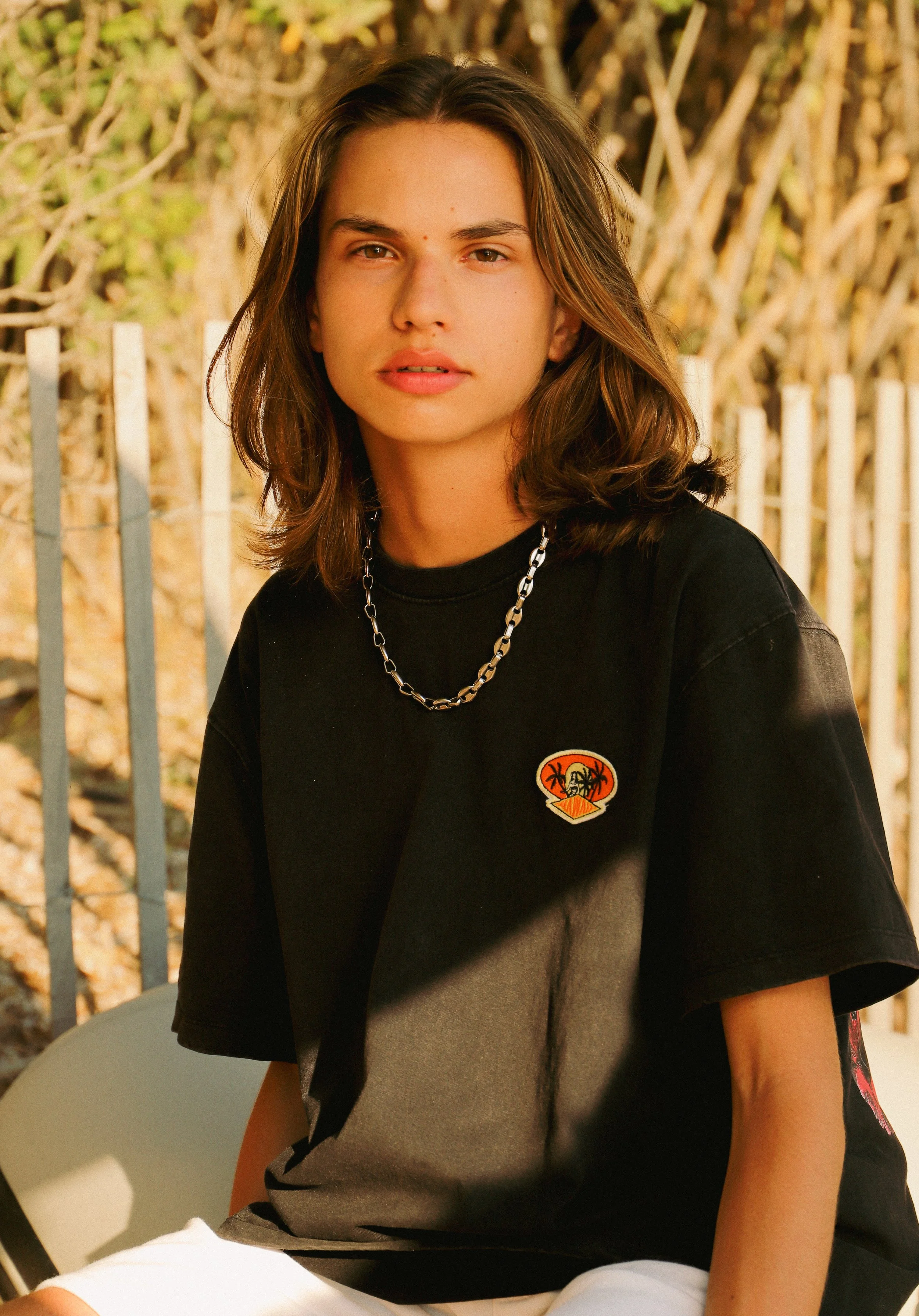 Jeune femme avec des cheveux bruns mi-longs, portant un t-shirt noir avec un patch coloré et un collier en chaîne, assise à l'extérieur contre une clôture en bois, en lumière naturelle.