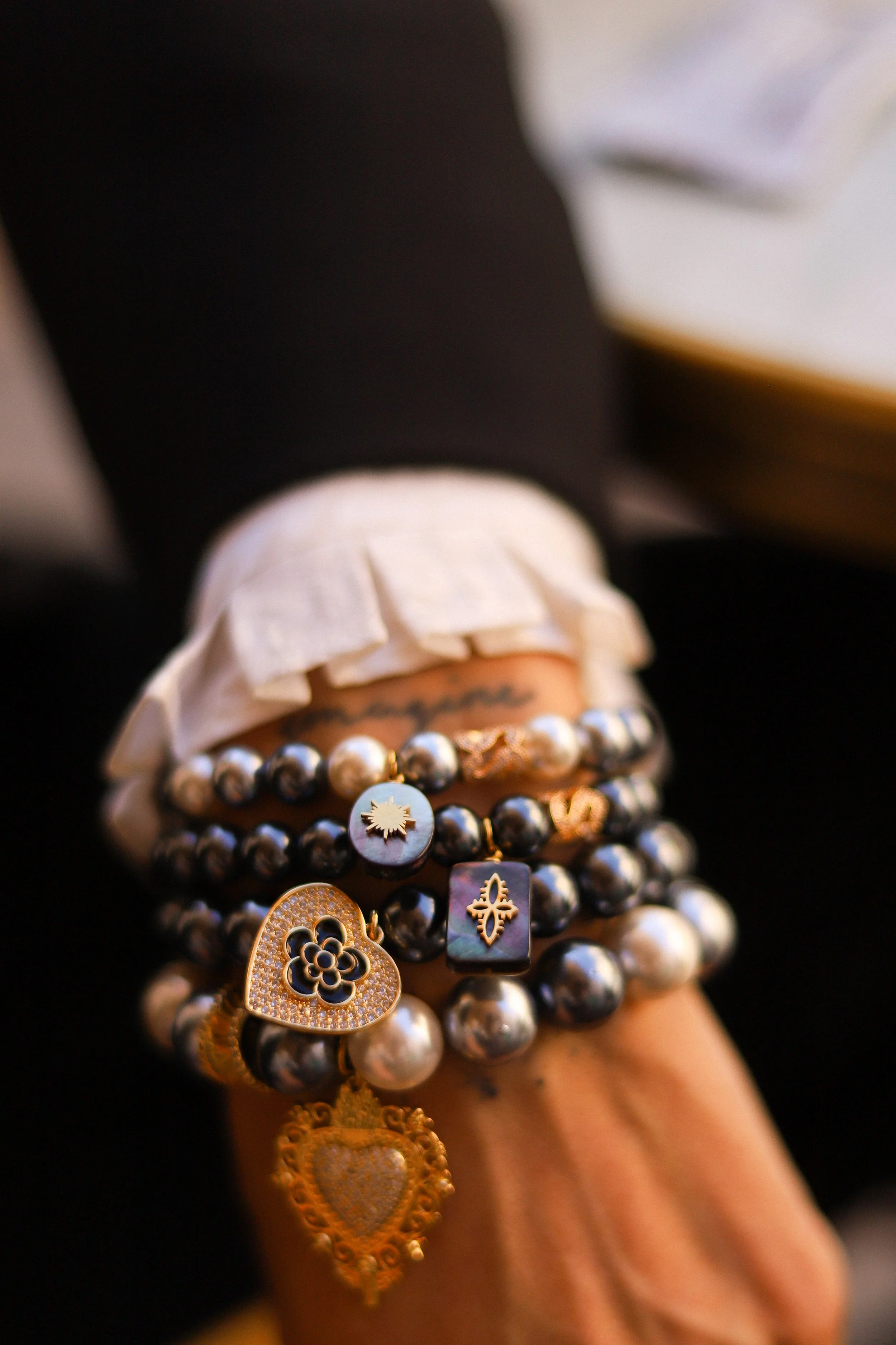 Poignet orné de plusieurs bracelets de perles de différentes couleurs et pendentifs, avec une main partiellement couverte d'une manche blanche et un vêtement noir en arrière-plan.