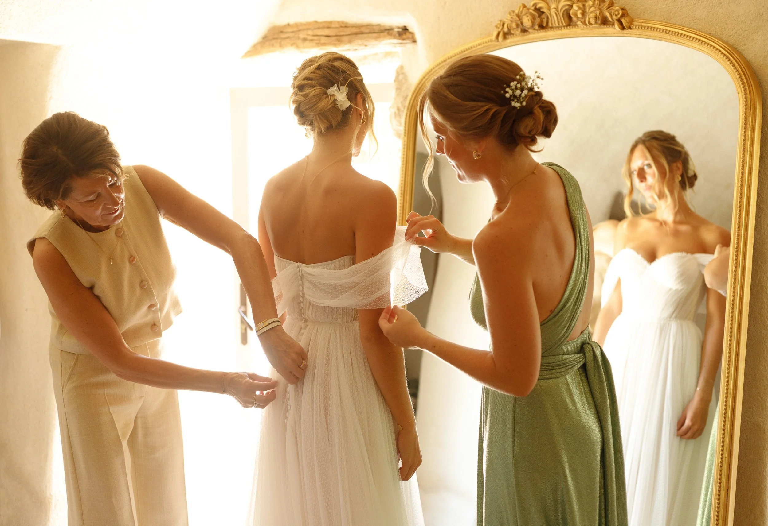 Une bride en robes blanches est aidée par deux femmes lors de la préparation de son vêtement dans une pièce lumineuse, avec un grand miroir ancien.