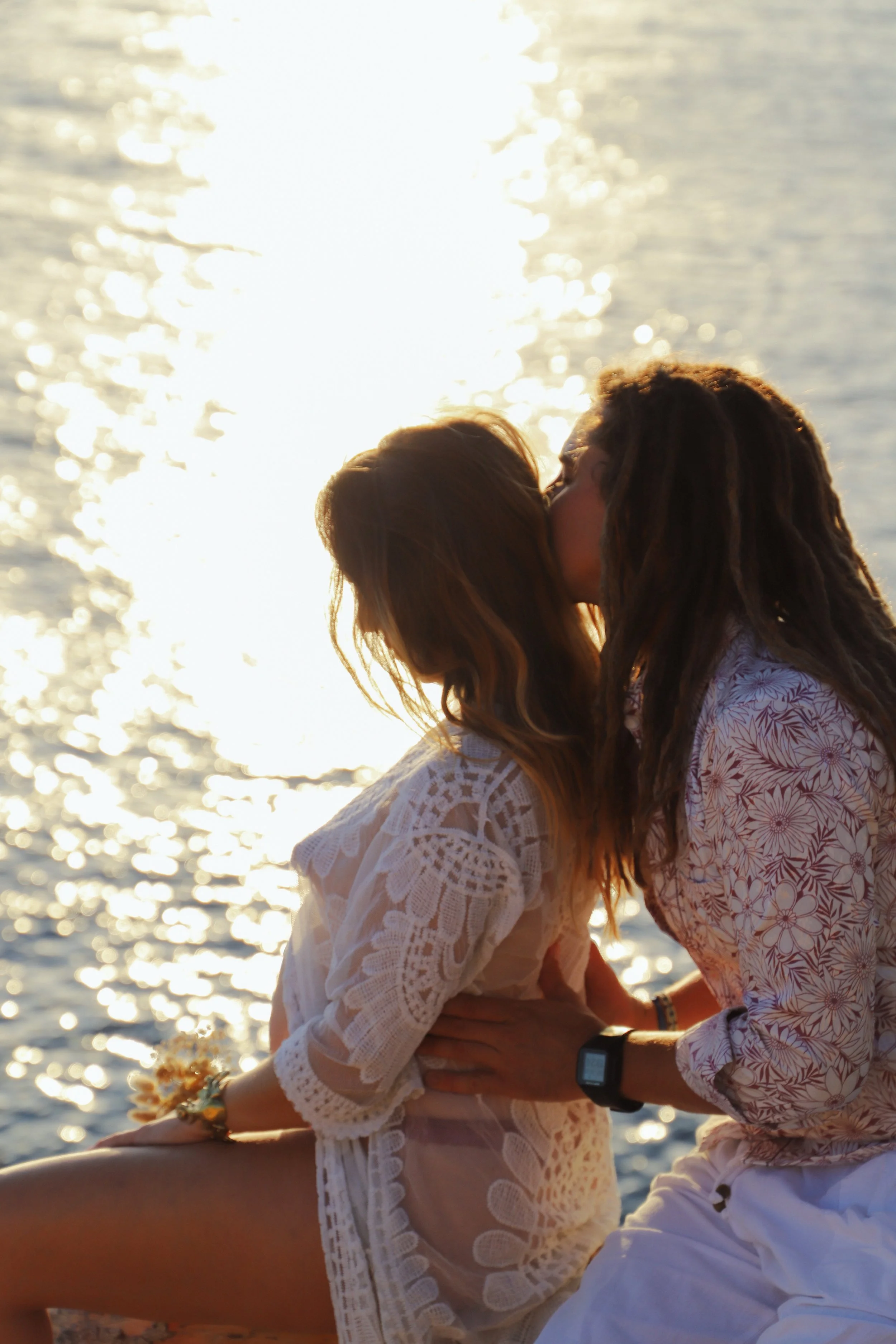 Deux femmes s'embrassent près d'un plan d'eau lors d'un coucher de soleil, vêtues de tenues légères et colorées.