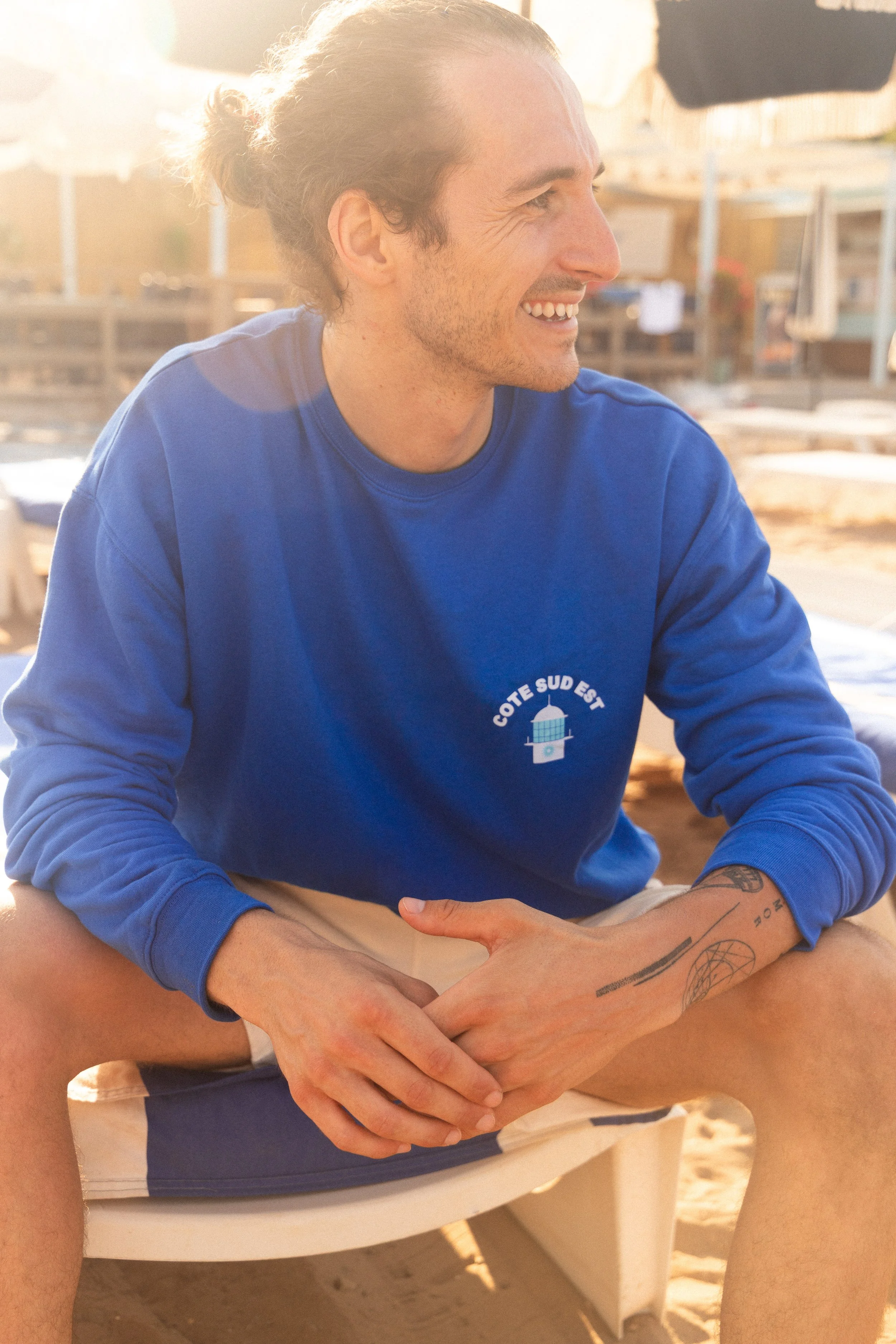 Un homme souriant assis sur une plage, portant un sweat bleu avec le texte 'CÔTE SUD EST' et un logo de phare.