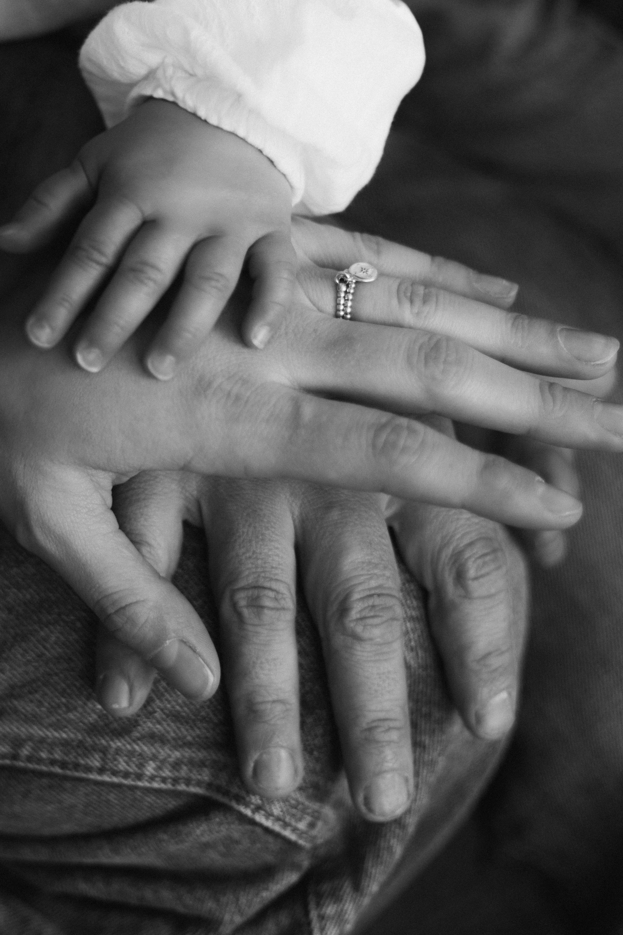 Une photo en noir et blanc de trois mains de tailles différentes, une main d'un bébé ou jeune enfant, une main d'une femme avec une bague, et une main d'un adulte, posées les unes sur les autres.