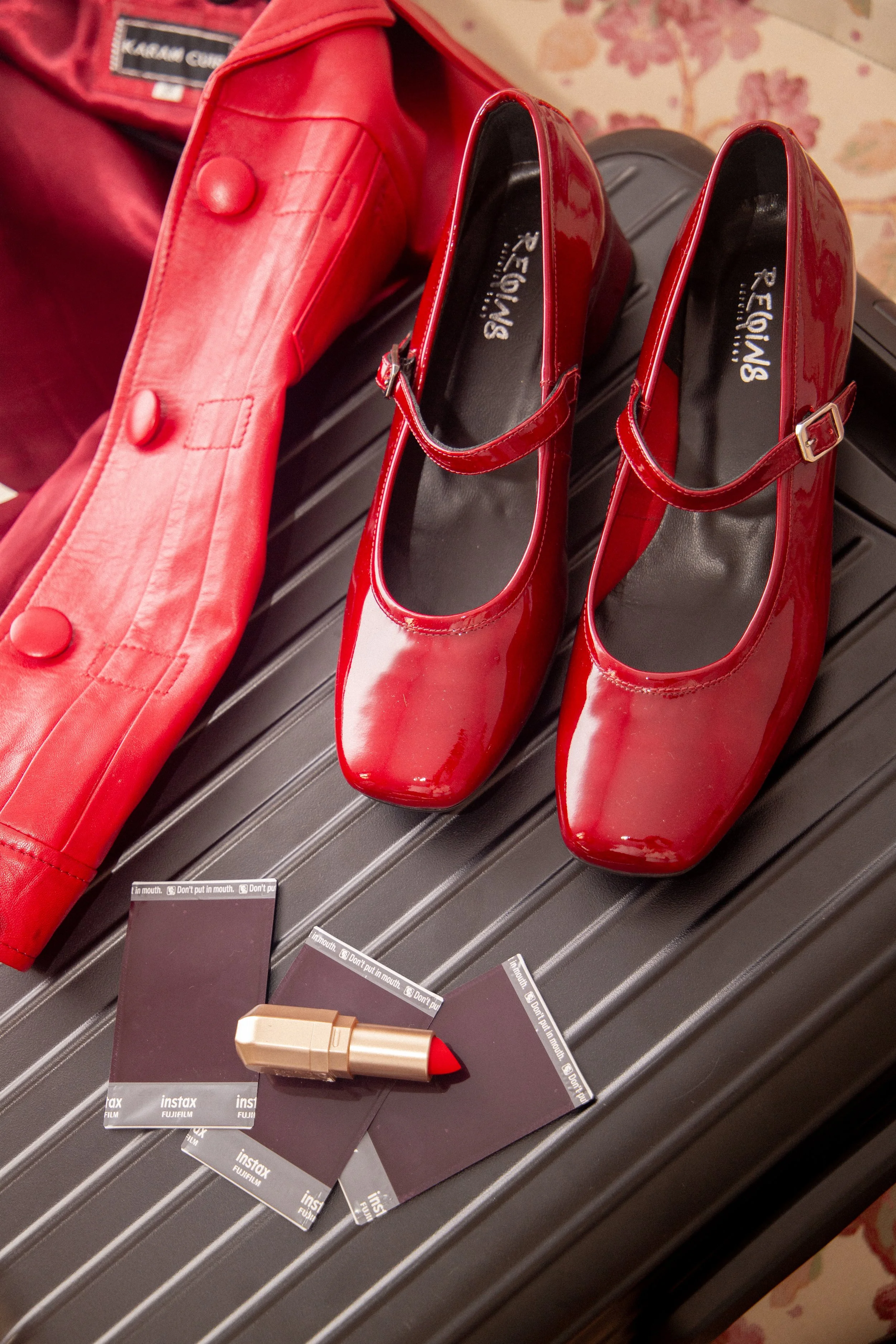 Une paire de chaussures à talons rouges, un manteau rouge en cuir, et trois cartes d'identité FujiFilm avec un rouge à lèvres rouge posé dessus.