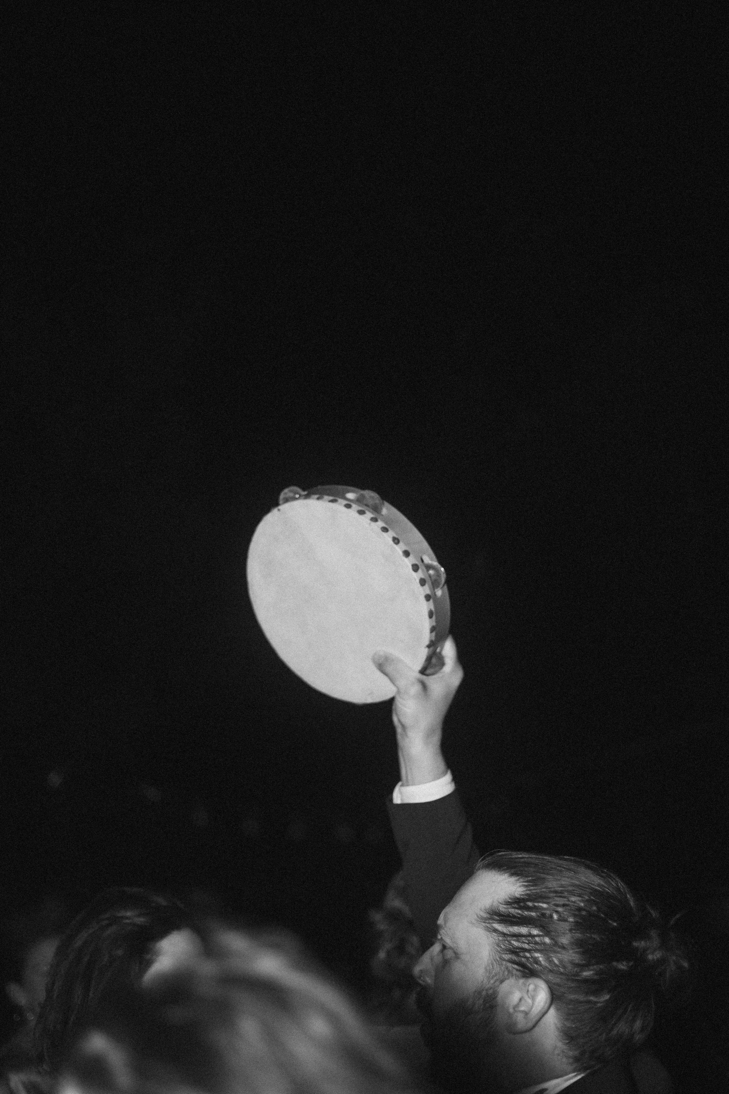 Une personne tient un tambour ou une tambourine dans une salle sombre, probablement lors d'un spectacle ou d'une performance. La personne a les cheveux bruns et une barbe, et porte un costume sombre.