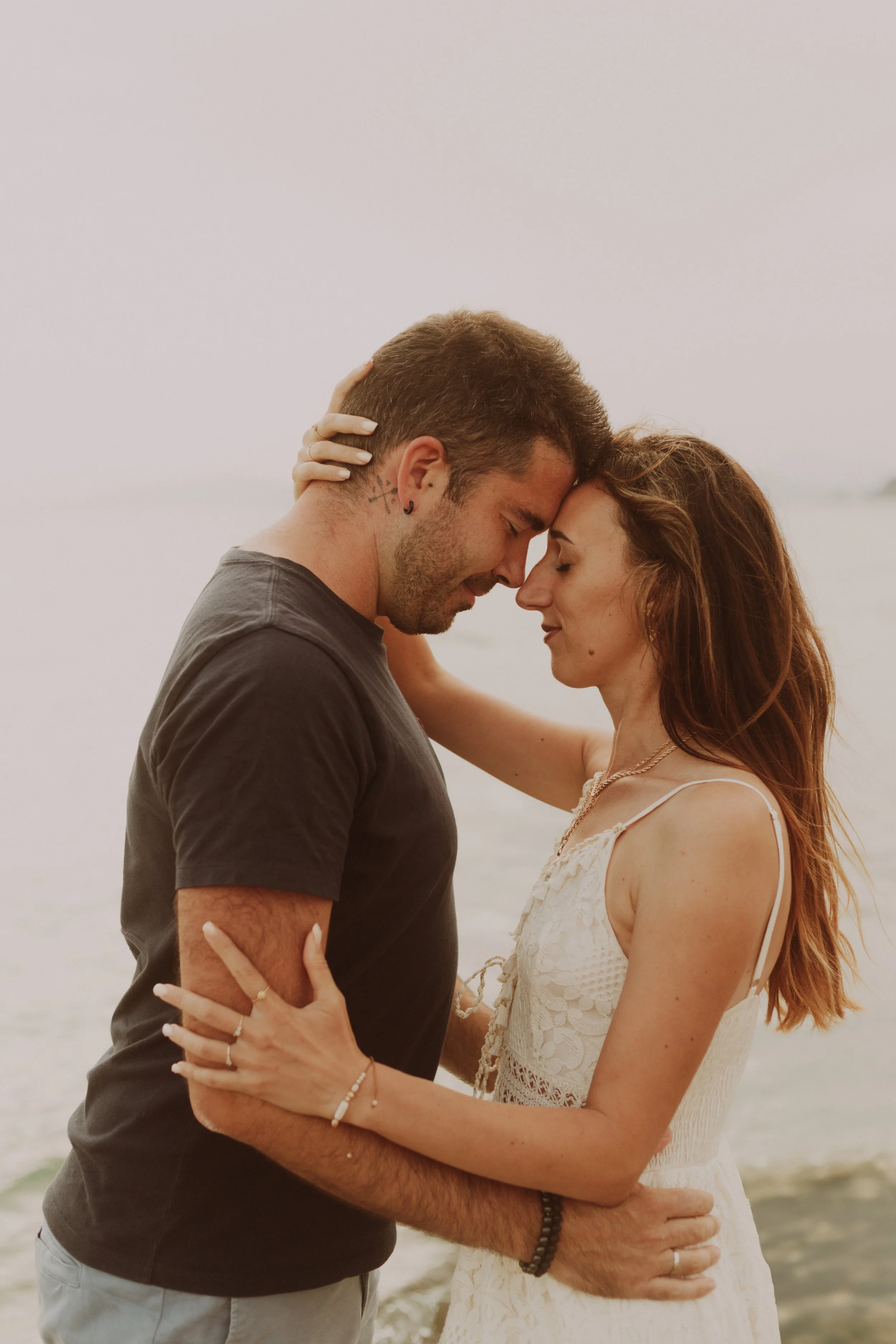 Un couple fusionnel se tient face à face sur la plage, avec la mer en arrière-plan, leurs fronts et nez touchant, créant une atmosphère intime et romantique.