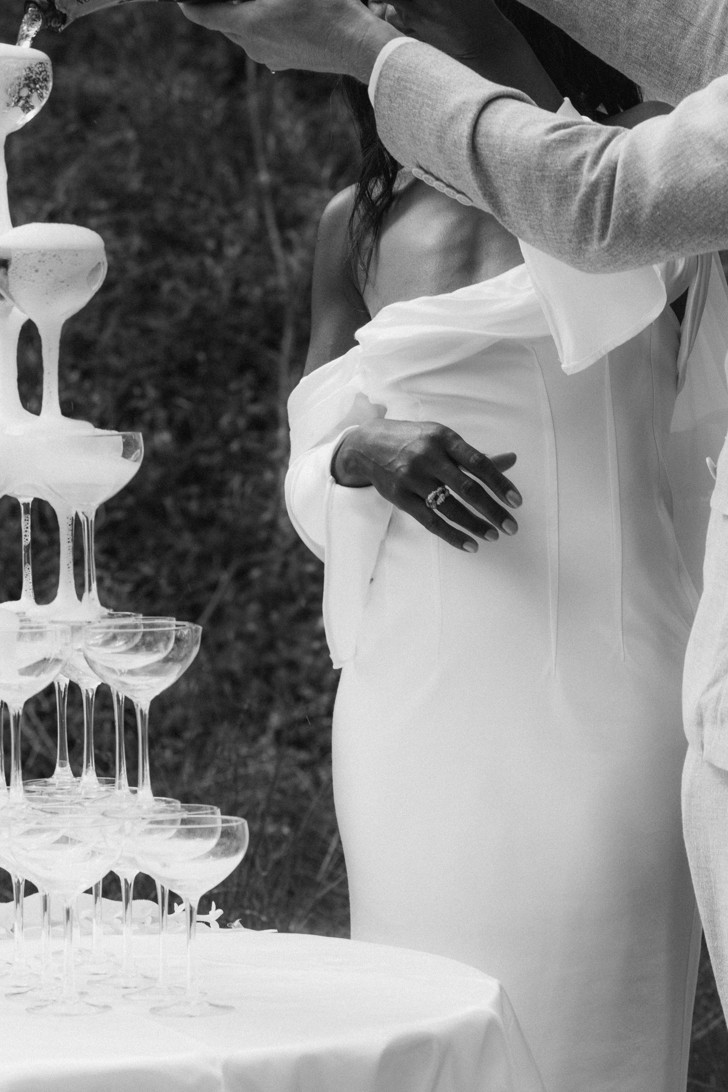 Une femme en robe blanche lors d'une célébration, avec un homme qui verse du champagne dans un tas de coupes à champagne sur une table.