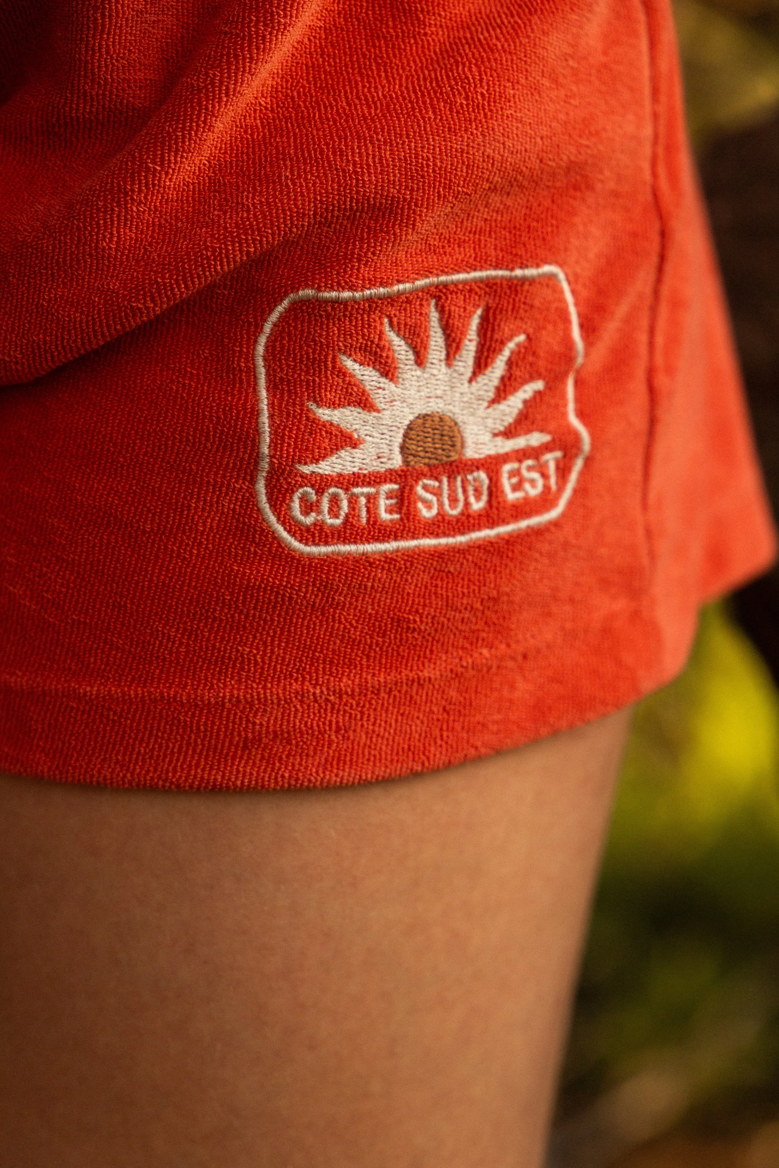 Détail d'un vêtement orange avec un patch brodé représentant un soleil, avec le texte 'COTE SUD EST'.