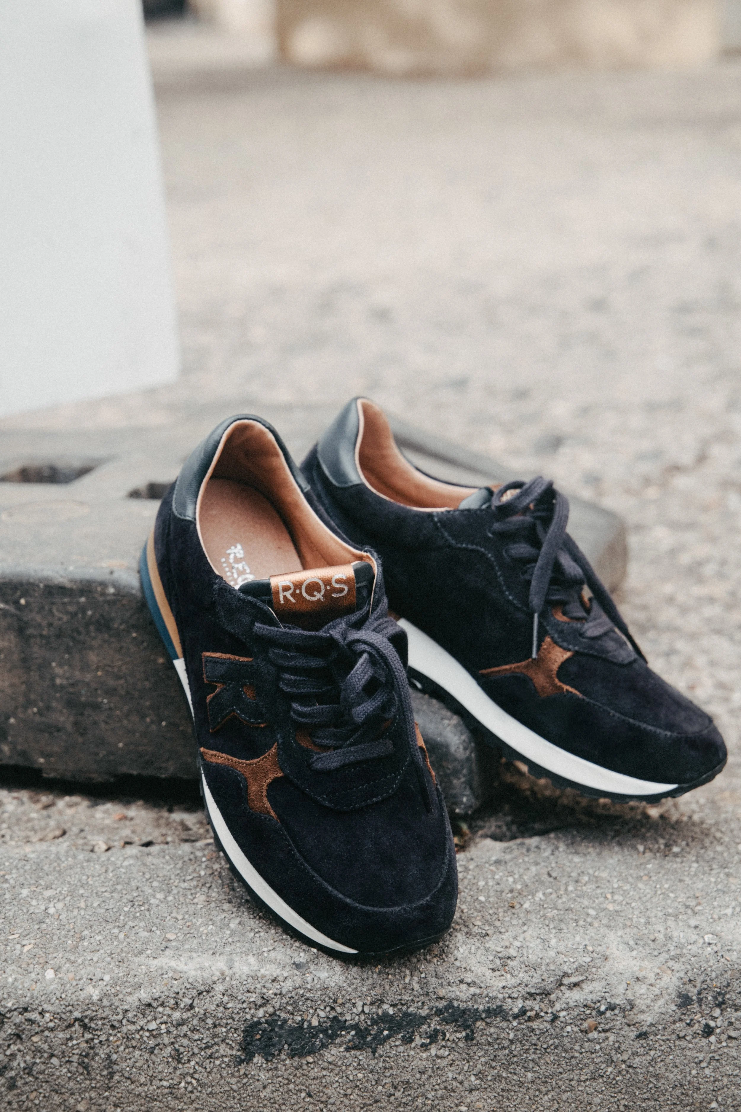 Une paire de baskets noires avec des accents marron, posée sur un trottoir en béton.