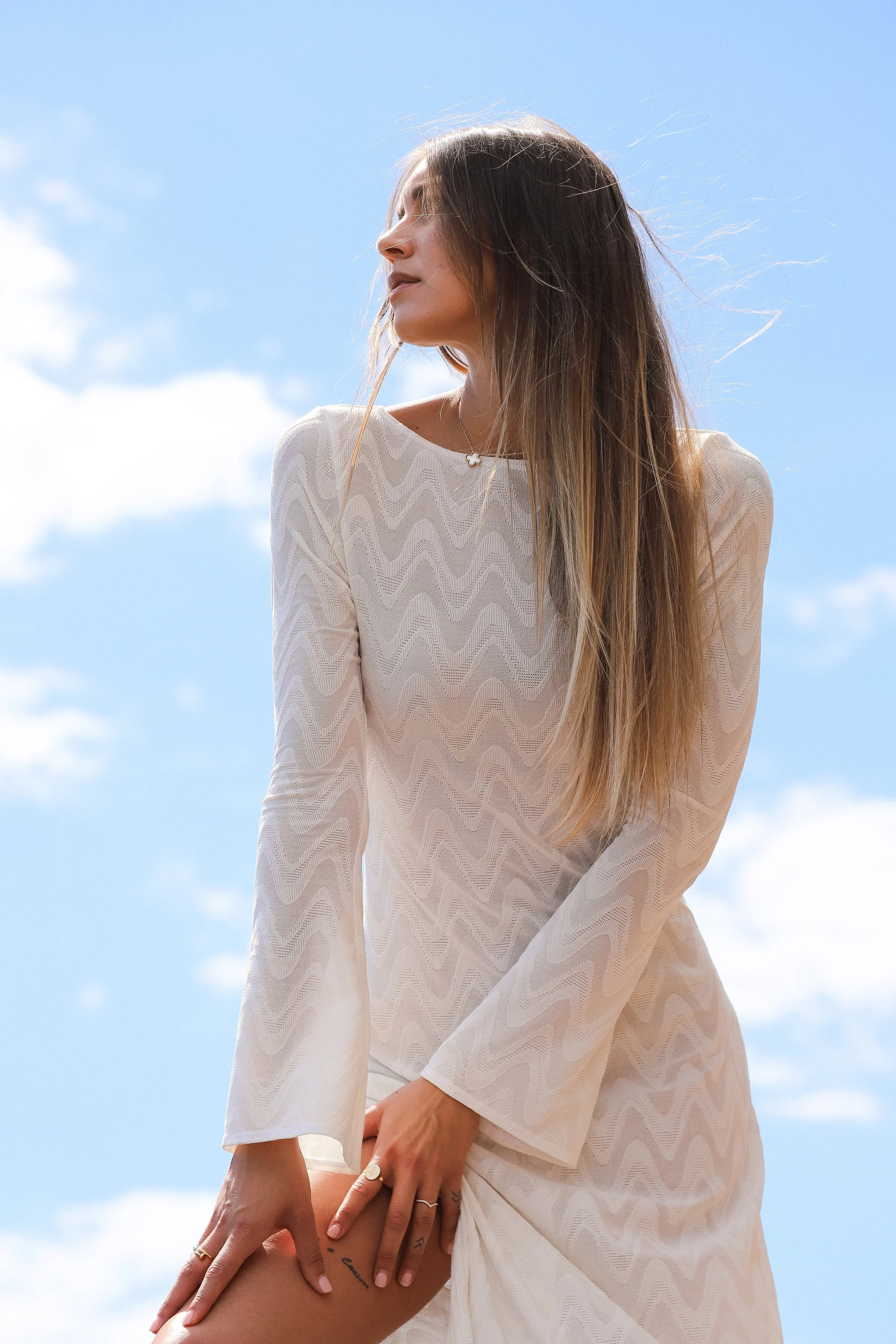 Jeune femme en robe blanche, debout sous un ciel bleu avec des nuages, regardant vers la gauche.