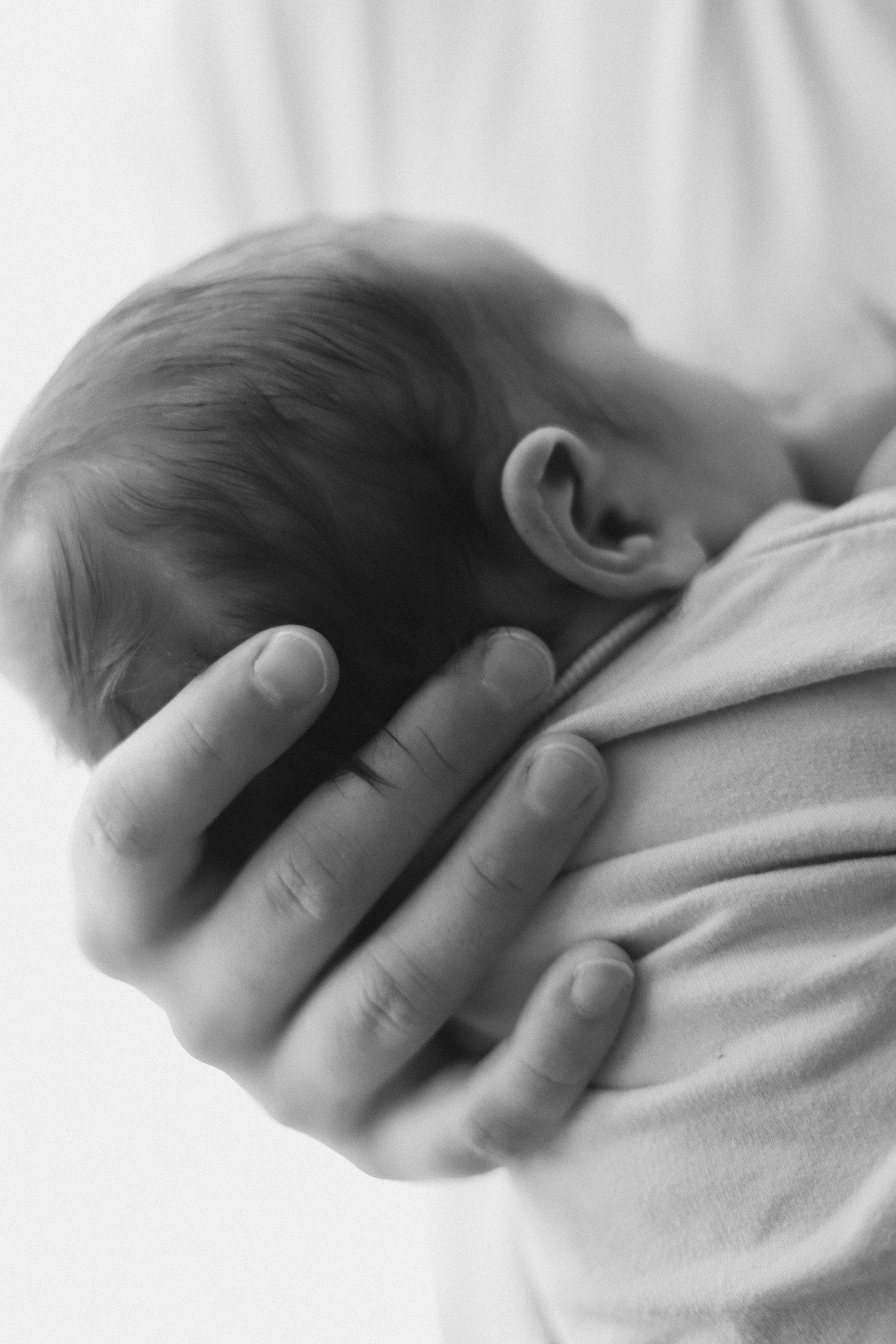 Une personne tient un bébé contre son épaule, le bébé a les cheveux foncés et porte un t-shirt clair, en noir et blanc.