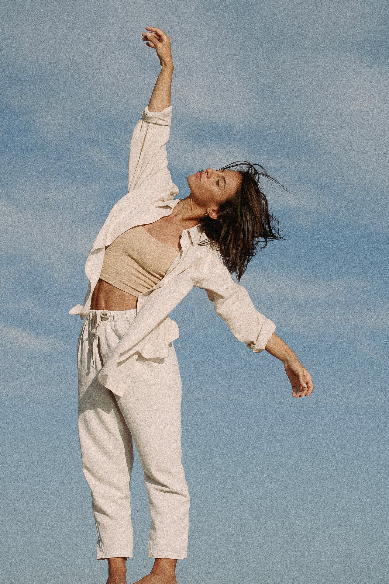 Une femme en tenue beige et blanche danse contre un ciel bleu, avec ses bras levés.