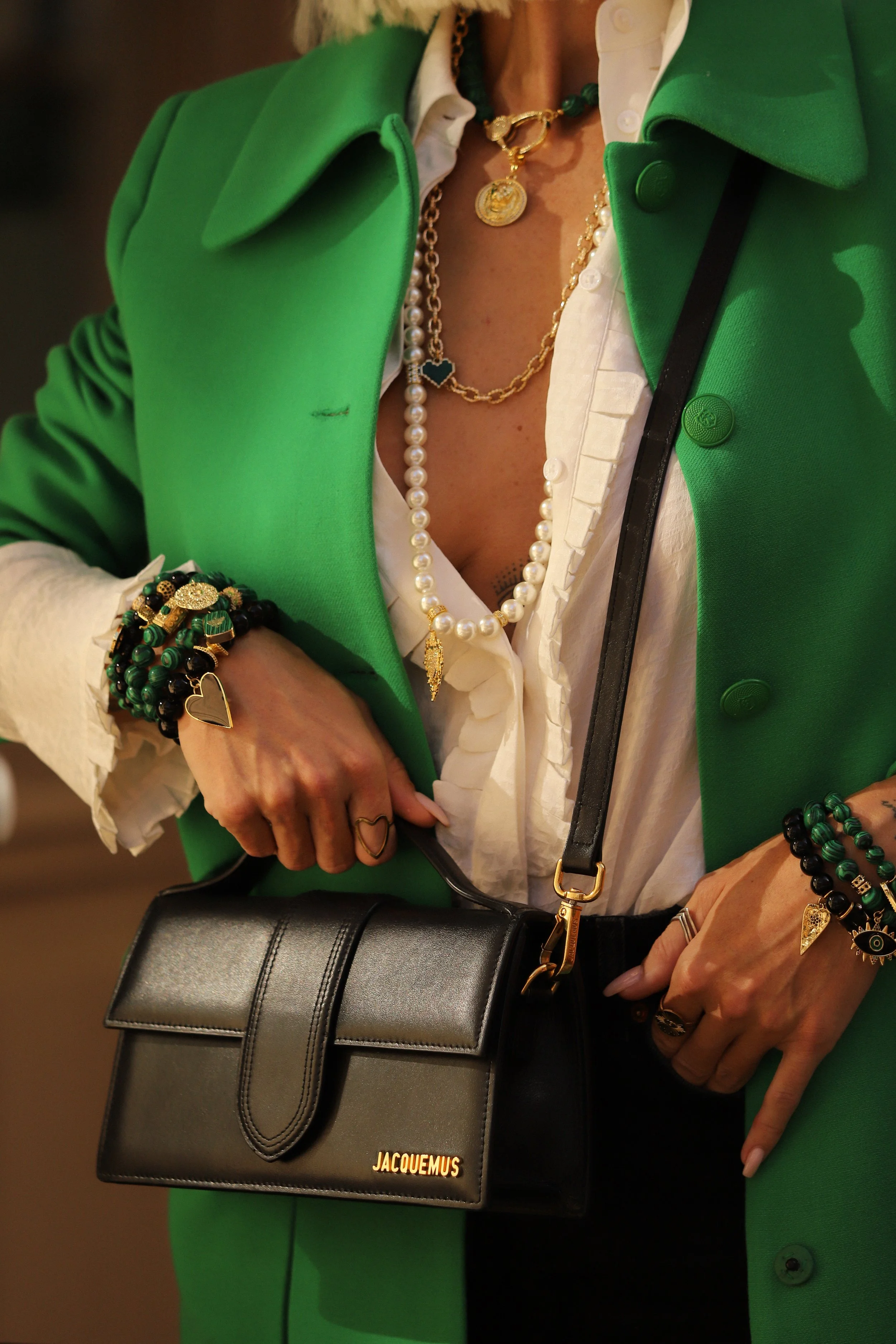 Une femme porte un trench vert, une chemise blanche, diverses colliers et bracelets, et tient un sac à main noir portant le nom Jacquemus.