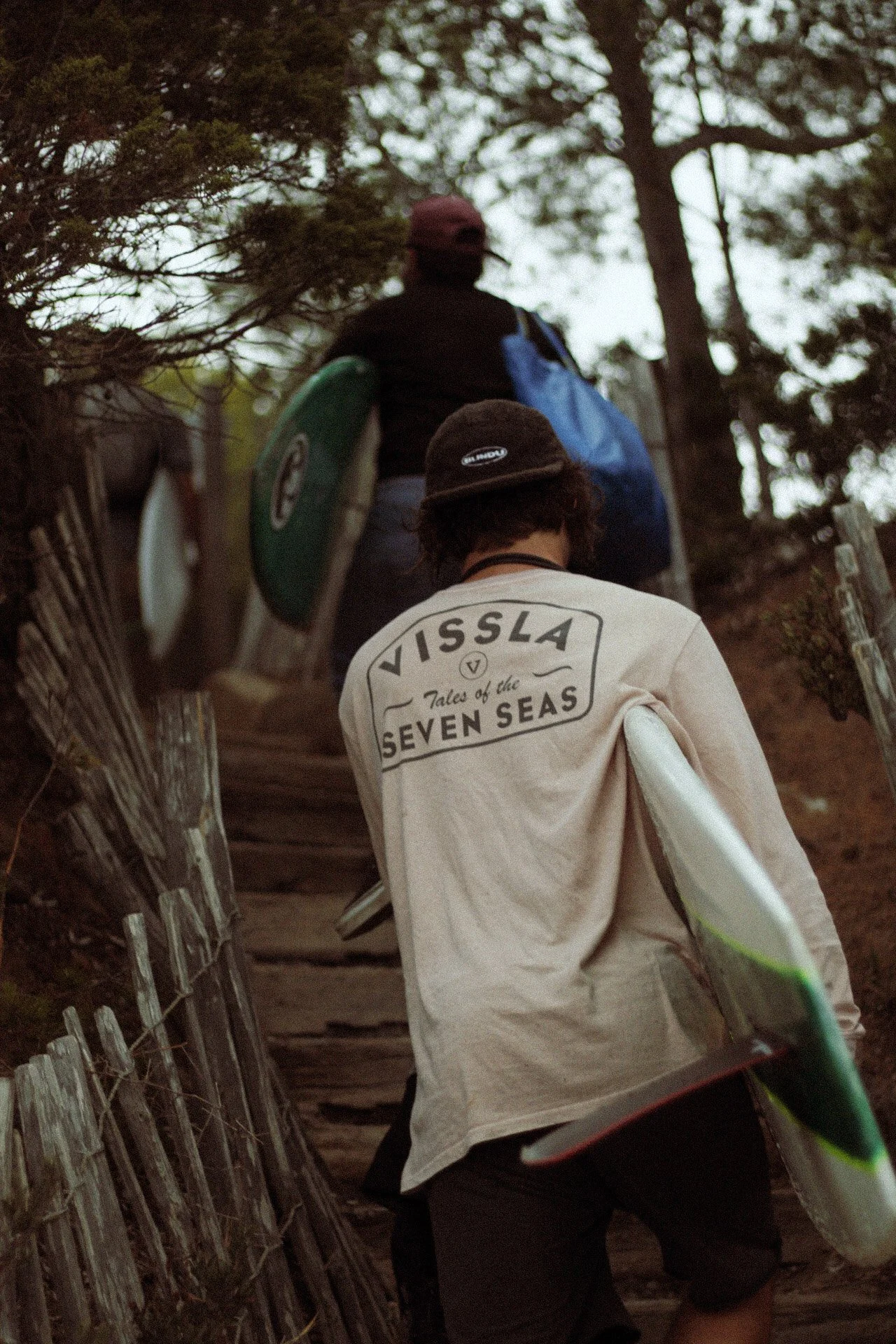 Deux personnes portant des planches de surf et un sac à dos montent un sentier en forêt avec des arbres autour.