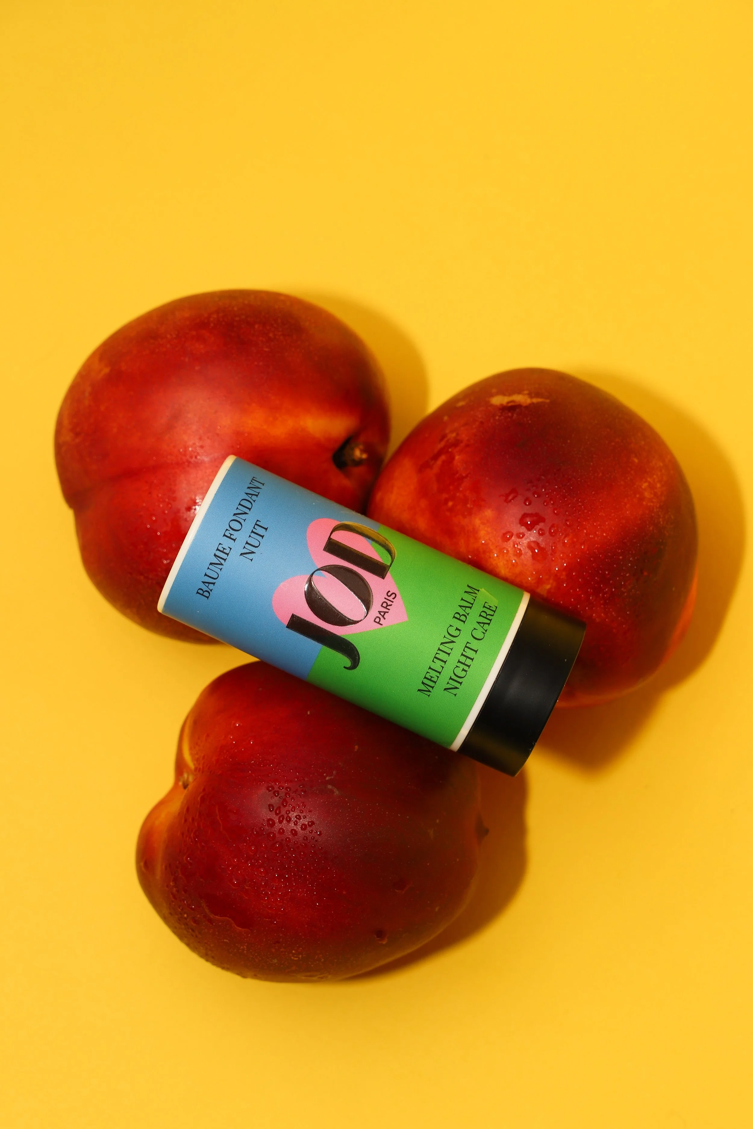 Trois pommes rouges avec un tube de baume à lèvres JOUÉ PARIS reposant sur elles, sur un fond jaune.