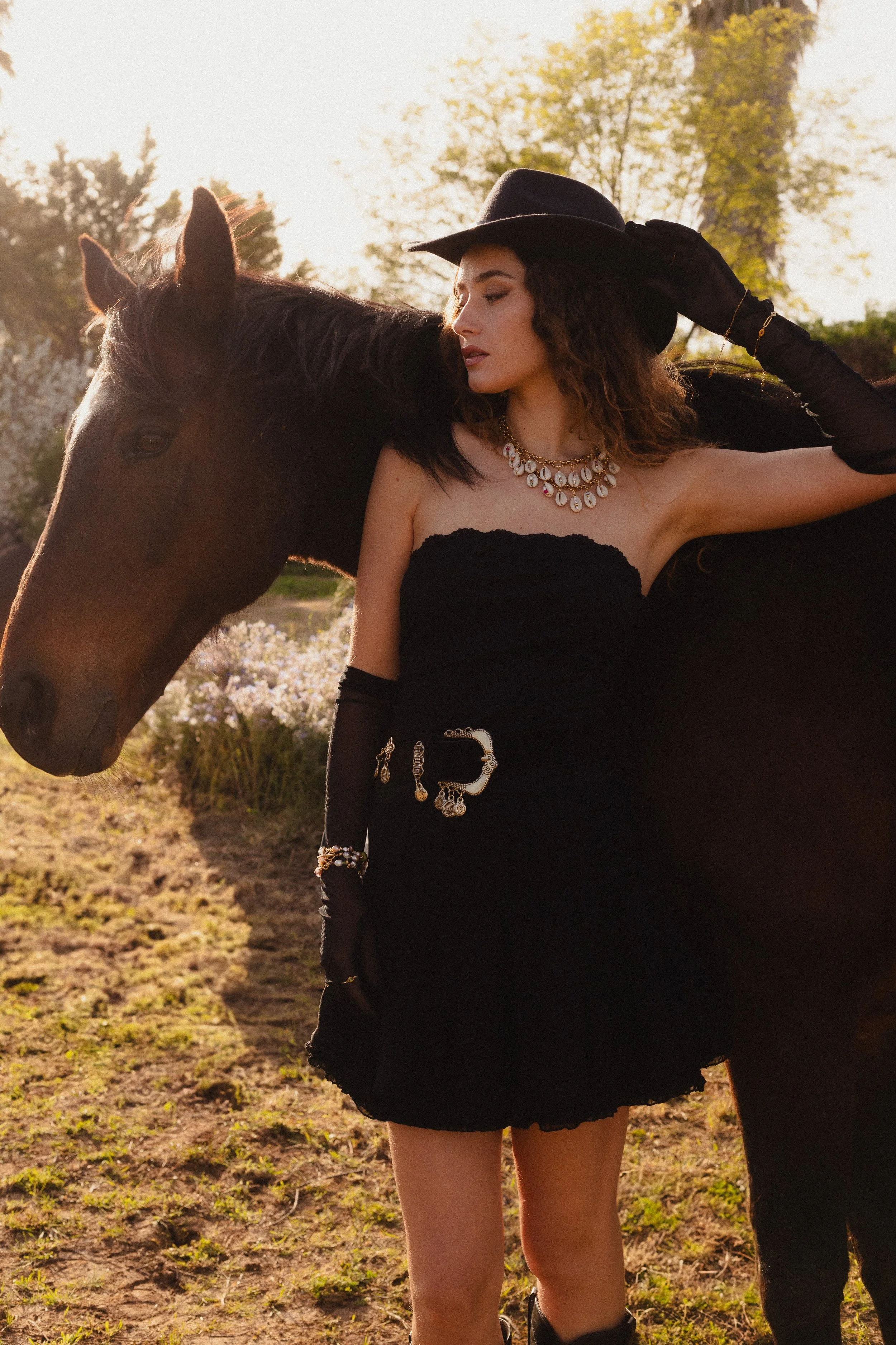 Une femme portant une robe noire, un chapeau noir, des bijoux élégants, et des gants longs noirs, pose à côté d'un cheval dans un champ ensoleillé avec des arbres en arrière-plan.