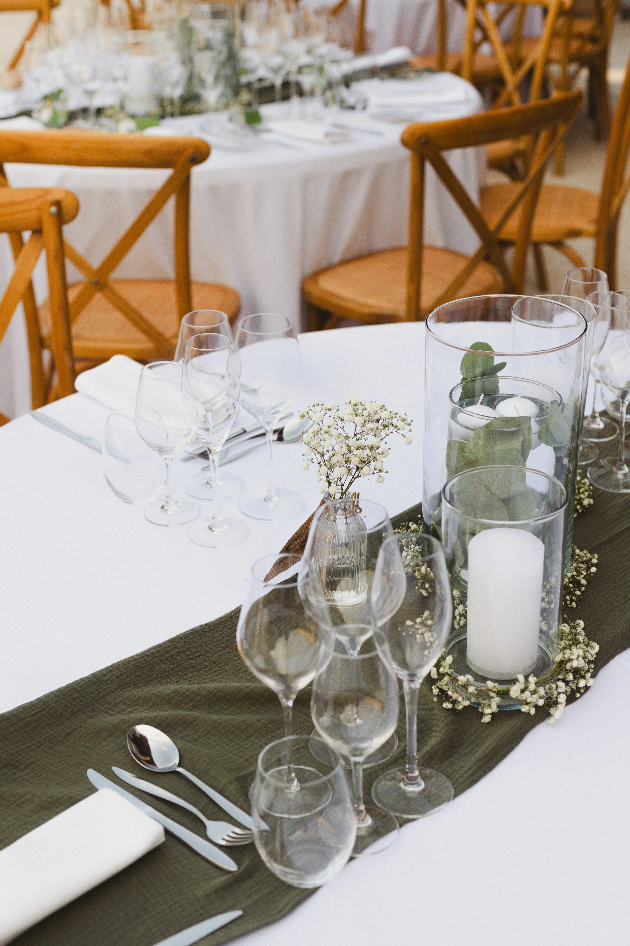Table de mariage avec décoration florale, bougies dans des cylindres en verre, carafe avec feuillage, et verrerie sophistiquée, entourée de chaises en bois.