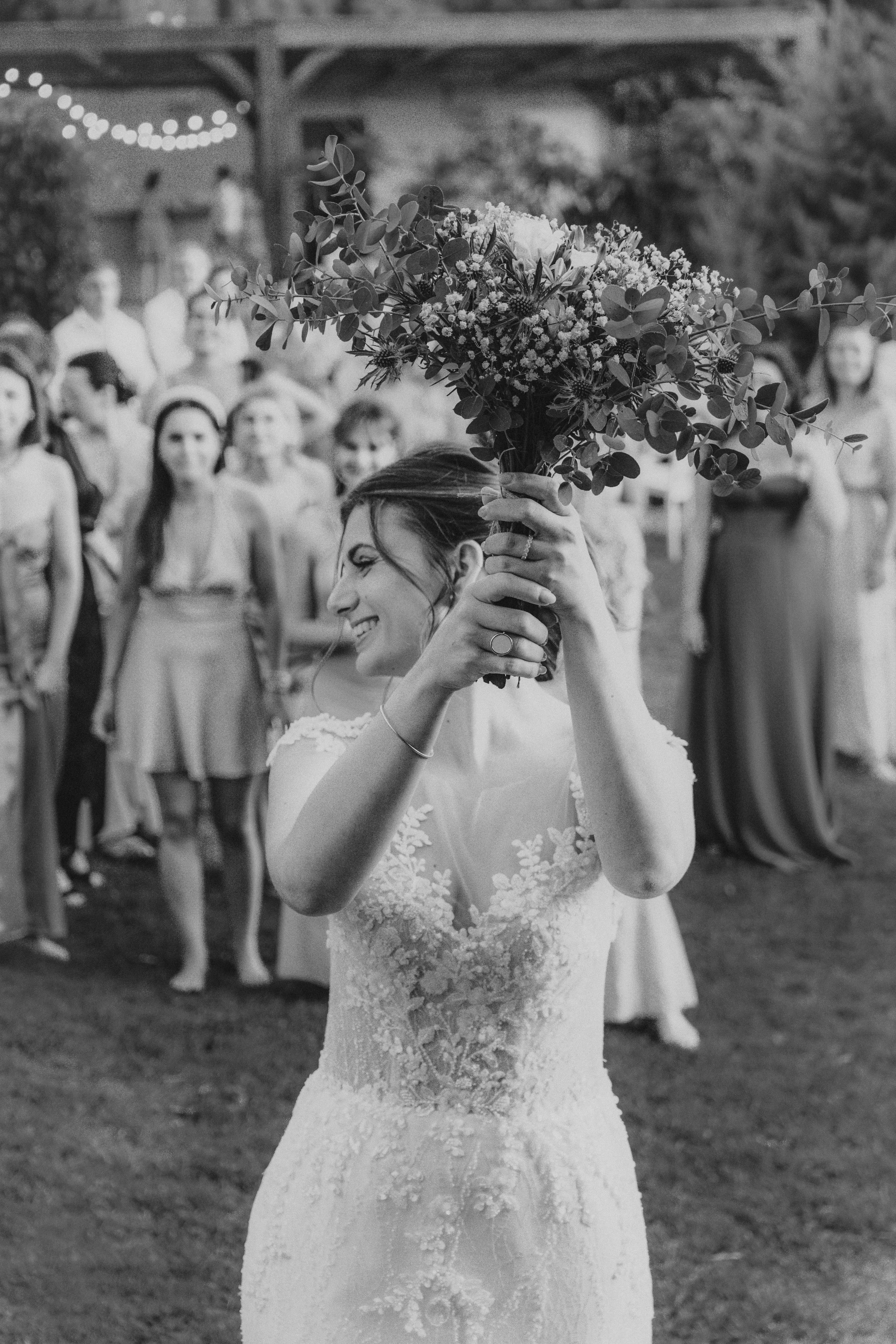Une femme en robe de mariage souriante avec un bouquet de fleurs, lors d'une cérémonie en plein air, entourée d'invités.