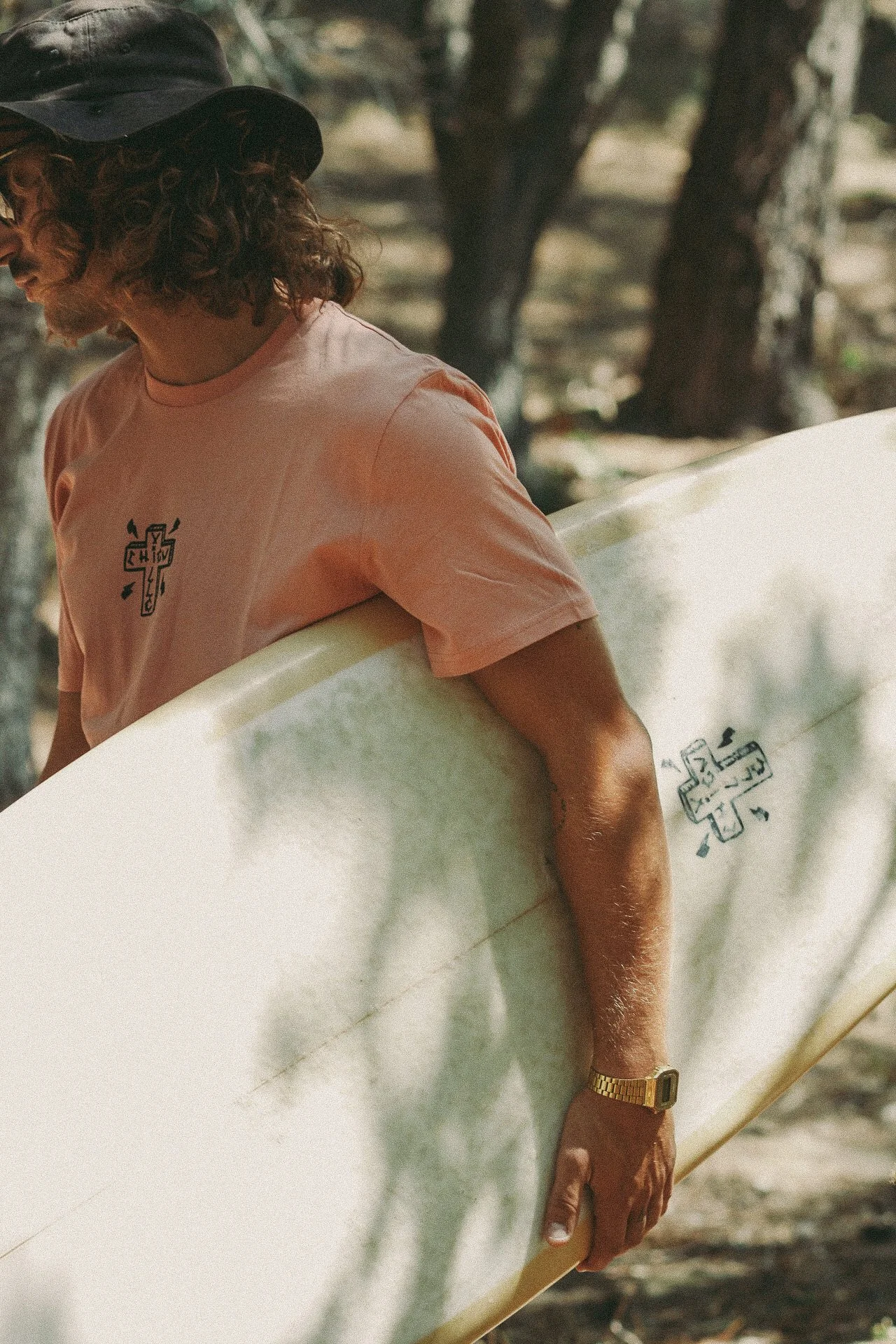 Un homme avec des cheveux bouclés, portant un chapeau noir, une chemise rose et une montre, tient une planche de surf dans un environnement boisé.