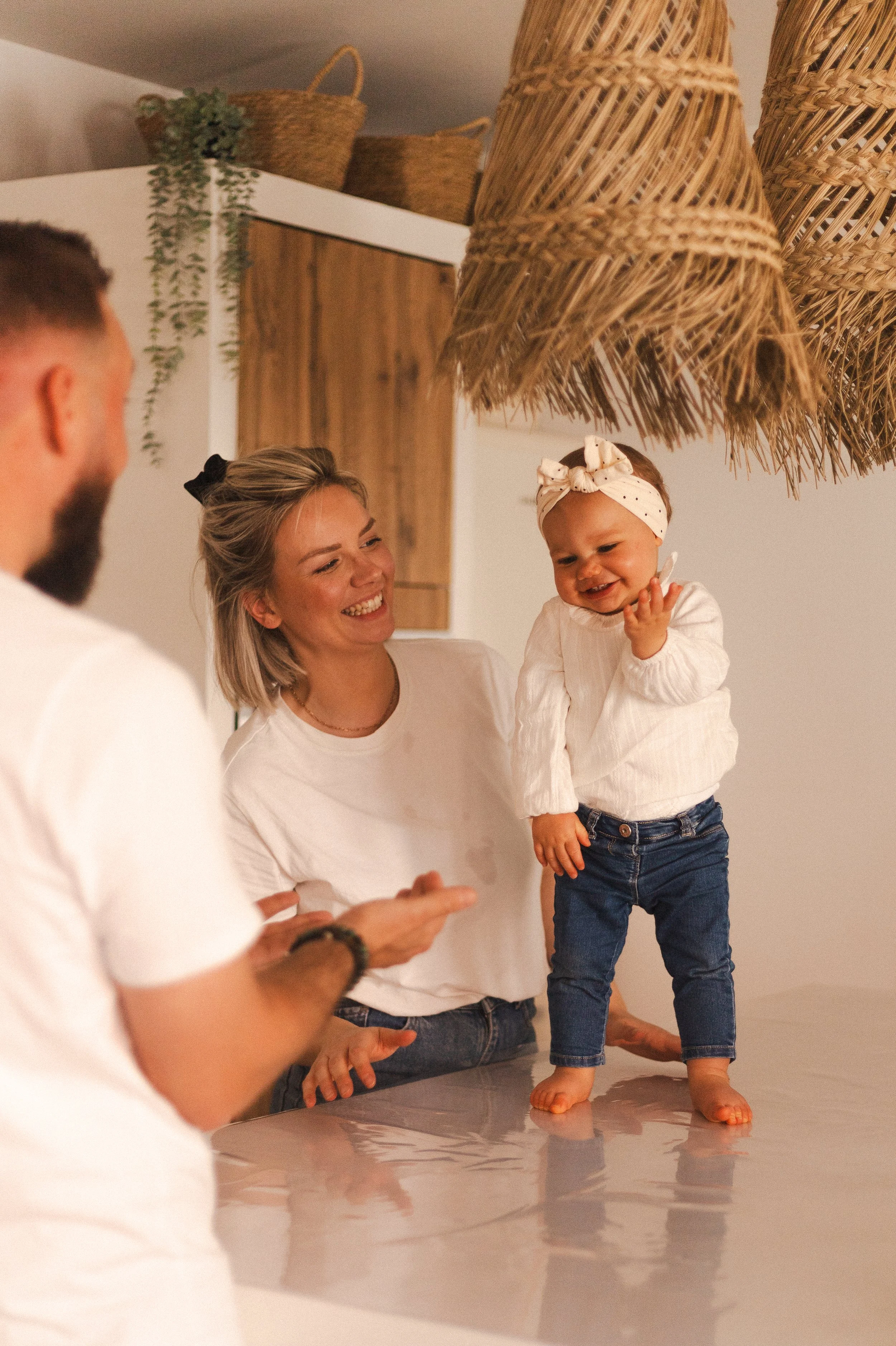 Une famille souriante dans une cuisine, avec une femme tenant un jeune enfant debout sur une table, portant un bandeau blanc avec des pois, et un homme en avant-apparence, en train de montrer quelque chose.